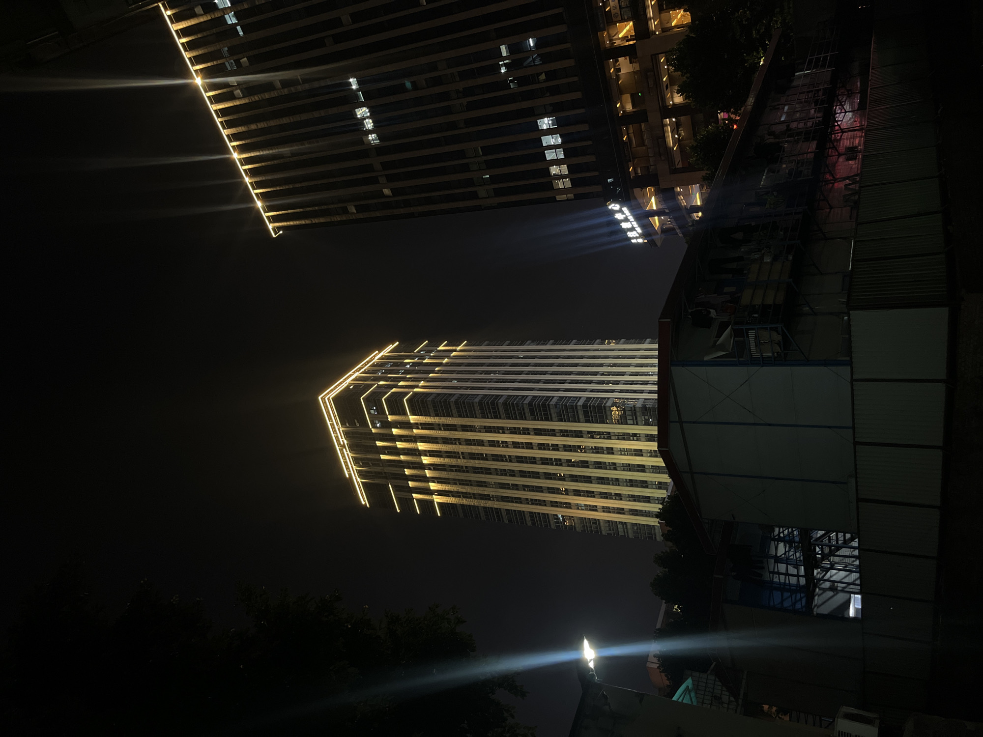 The image depicts a nighttime urban landscape in Yuzhong District, China, characterized by prominent illuminated high-rise buildings and foreground structures. On the left, a tall, slender skyscraper features extensive yellow-golden LED strip lighting outlining its top perimeter and running vertically along its facade, illuminating numerous windows within. To the right, another, slightly taller skyscraper stands with a darker, vertically textured facade, also displaying golden illumination along its top edge. Its lower sections reveal multiple lit windows and a multi-story commercial or retail base, with discernible Chinese characters on a sign located on a lower floor.
In the mid-ground, below the skyscrapers, are several low-rise, modular-looking structures with light-colored panels and dark roofs, enclosed by a corrugated metal fence. These structures appear to be temporary buildings, possibly construction site offices or dormitories, and exhibit minimal, internal illumination. Dark tree foliage is visible on the far left and also in the mid-ground between the foreground structures and the main buildings. A bright, blue-white streetlamp on a pole is positioned on the left side of the frame, emitting a strong upward light beam. The sky is dark, indicating a clear night. No individuals are visible within the scene.