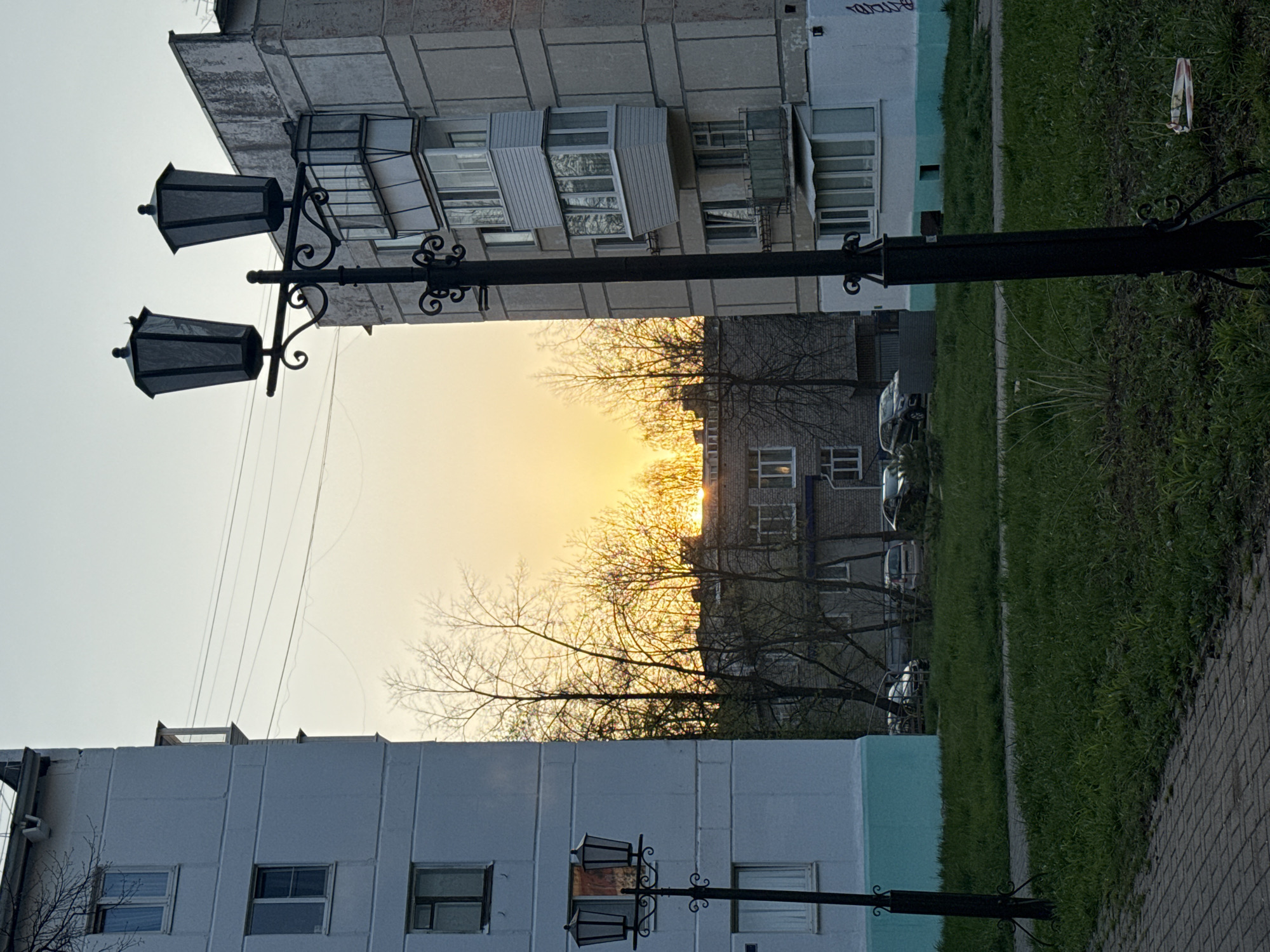 The image shows an urban scene in what is presumed to be Chaikovsky, Russia, given the prompt. The photograph is taken at ground level, looking upwards towards a street scene framed by buildings on either side. In the foreground, a black, ornate lamppost stands on both sides of the image, partially obscuring the view. Between the buildings, the setting sun casts a golden glow, with silhouetted tree branches adding detail to the skyline. Buildings on either side show signs of wear and tear, indicative of older construction. A grassy area runs along the right side of the frame, beside a paved walkway. Parked cars are visible between the buildings in the background. The sky is visible in the upper part of the image, with a light, pale blue hue. There are no people visible in the scene. The overall mood is serene, capturing a quiet moment in the city at sunset.