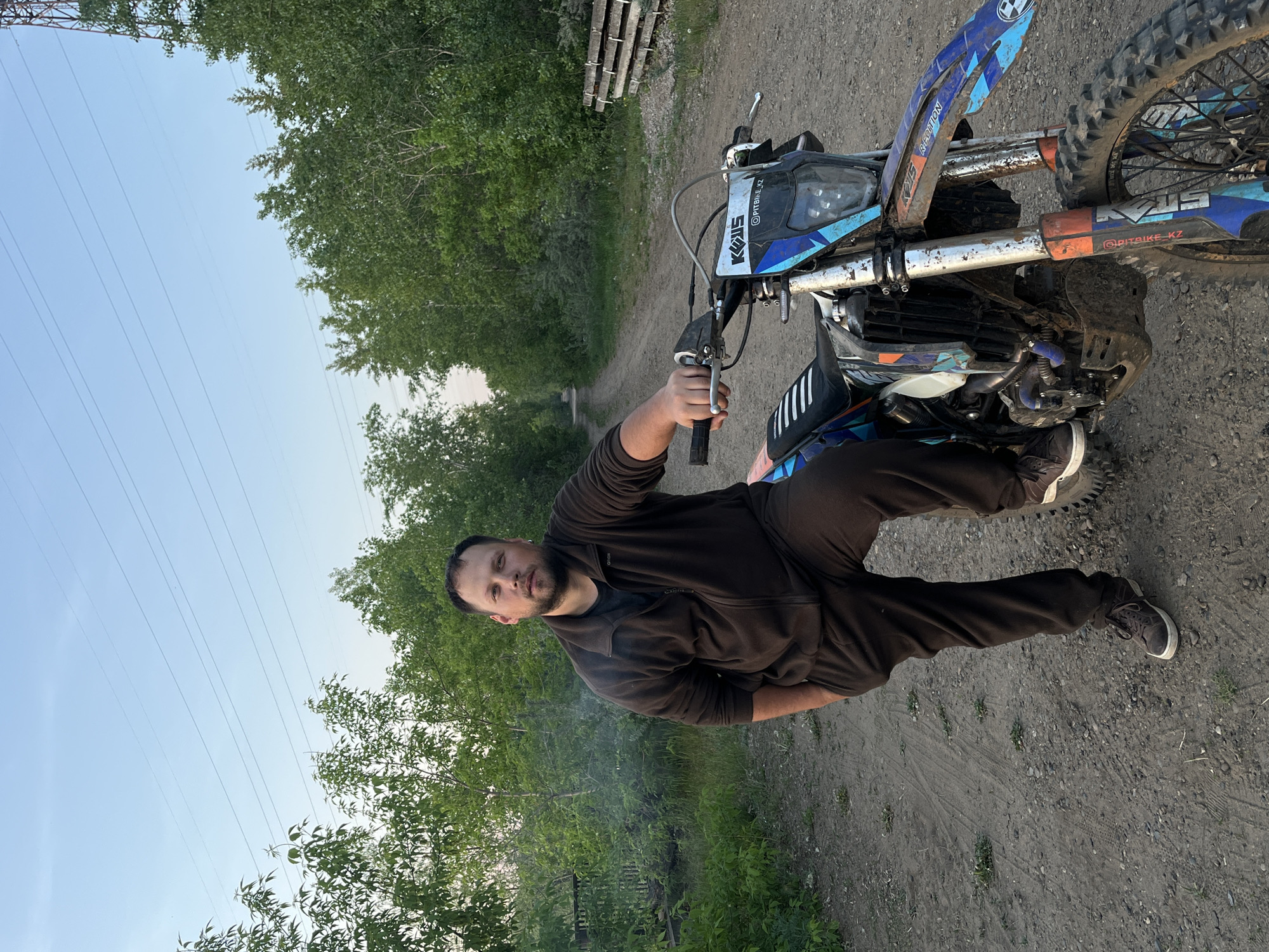 A man stands alongside a dirt bike on a dirt road. He is wearing a brown track suit and dark sneakers. The dirt bike is blue and black. Behind the man are trees and some power lines. The location is outdoors in a natural setting, likely near Chaikovsky, Russia.