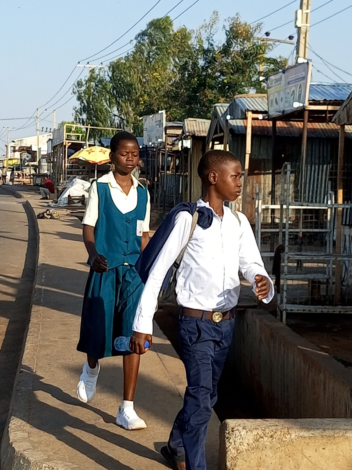 In Garko, Nigeria, two young people walk along a paved path adjacent to a road lined with market stalls and utility poles. The individual on the left, appearing to be a girl, wears a school uniform consisting of a light-colored shirt, a teal vest, and a matching teal skirt. She carries a clear plastic water bottle in her right hand. The person on the right, seemingly a boy, is dressed in a white long-sleeved shirt, dark blue trousers, and a brown belt with a circular buckle. He has a dark jacket draped over his left shoulder. The market stalls are constructed with corrugated metal roofs and sides, and the ground is a mix of concrete and dirt. Trees with dense foliage are visible in the background. The scene is illuminated by bright sunlight, casting distinct shadows on the ground.