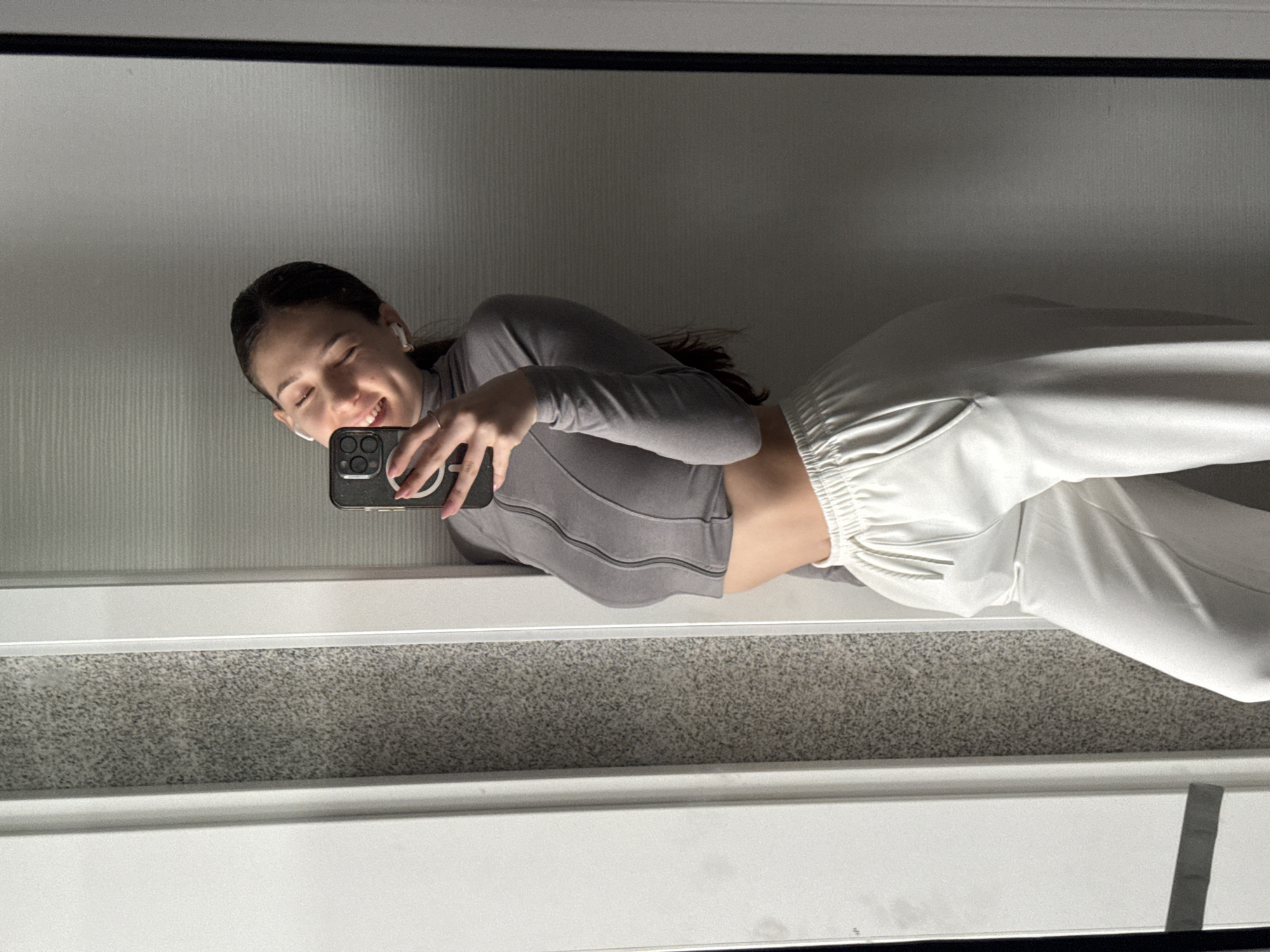 An image displays a young woman taking a mirror selfie. She is captured sideways in the reflection, with her head towards the left and her body extending towards the right. She has dark hair and is wearing white wireless earbuds. A wide, joyful smile spreads across her face, and her eyes are squinted with happiness.
She is dressed in a form-fitting, long-sleeved grey crop top featuring horizontal ribbed textures, which reveals a section of her midriff. Her lower body is clad in loose-fitting, light-colored (likely white or off-white) sweatpants or joggers with an elasticated waistband and cuffs. She holds a dark-colored smartphone, likely an iPhone given the camera module design, in her right hand, positioning it to capture her reflection.
The background visible in the mirror behind her is a light-colored wall with subtle vertical striations. Below the reflective surface, there is a speckled grey texture, possibly a carpet or floor, bordered by clean white framing elements. The overall setting appears to be an indoor space with good illumination, suggesting a casual and relaxed atmosphere. No visible text or distinctive location cues are present.