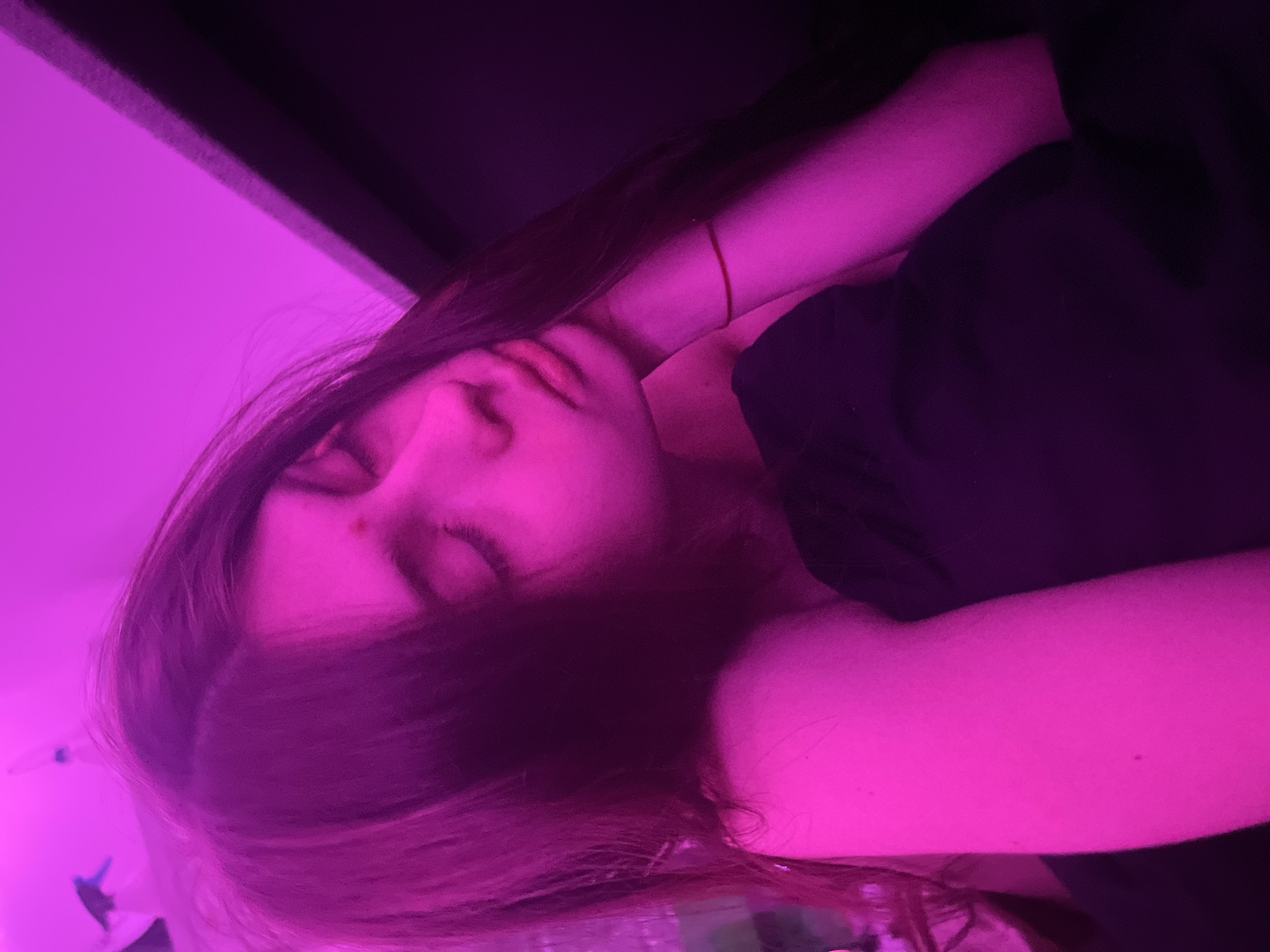 The image presents a close-up portrait of a young person, likely female, with long dark hair, resting or sleeping in a dimly lit indoor setting. Their eyes are closed, and their head is tilted to one side. A thin red string bracelet is visible on their raised right wrist. A small reddish mark is noticeable on the bridge of their nose.
The scene is dramatically illuminated by a strong, vibrant magenta light source, which casts a striking purple and pink hue across the subject's skin, hair, and surroundings. This artificial lighting creates a moody and ethereal atmosphere. The person is wearing a dark, possibly black, top or shirt, which appears darker under the intense colored light.
In the background, a brightly lit magenta wall is visible on the left, contrasting with darker, shadowed areas that suggest a wall or ceiling in the upper right. The overall impression is one of peace and serenity, capturing an intimate and quiet moment. The absence of natural light suggests it is evening or night. There is no visible text in the image. This scene is set in Saint Petersburg, Russia.