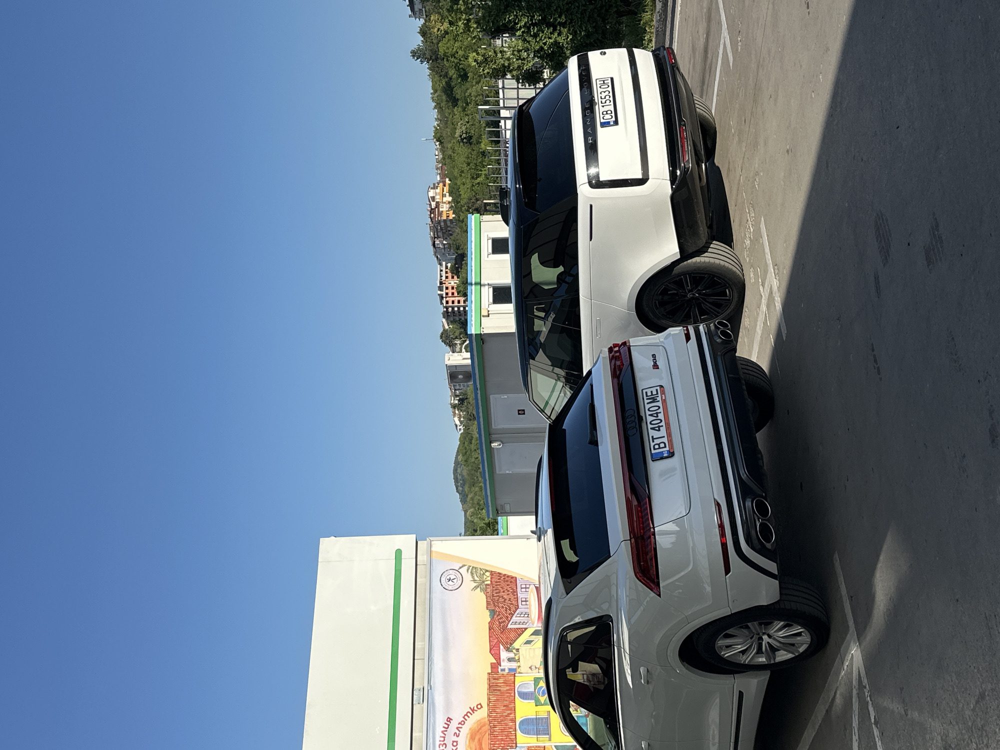 The image depicts a sunny day in Veliko Tarnovo, Bulgaria, with two white SUVs parked in a lot. The sky is a clear, vibrant blue, indicating fair weather and likely mid-morning or early afternoon. The foreground shows the rear of an Audi SUV, identifiable by its taillights and the license plate "BT 4040 ME". Behind it, a white Range Rover is parked, with its license plate visible as "CB 1553 OH". Both vehicles are modern and appear to be in good condition.
In the background, there is a building with colorful murals, possibly indicating a commercial or recreational area. Beyond the building, a hillside is covered with lush green trees and a few residential buildings with distinct architecture, characteristic of the region. The parking lot is paved, with visible white lines marking the parking spaces, and a section is in shadow, suggesting the sun is casting strong shadows. The overall scene is peaceful and captures a moment of everyday life in a Bulgarian town.