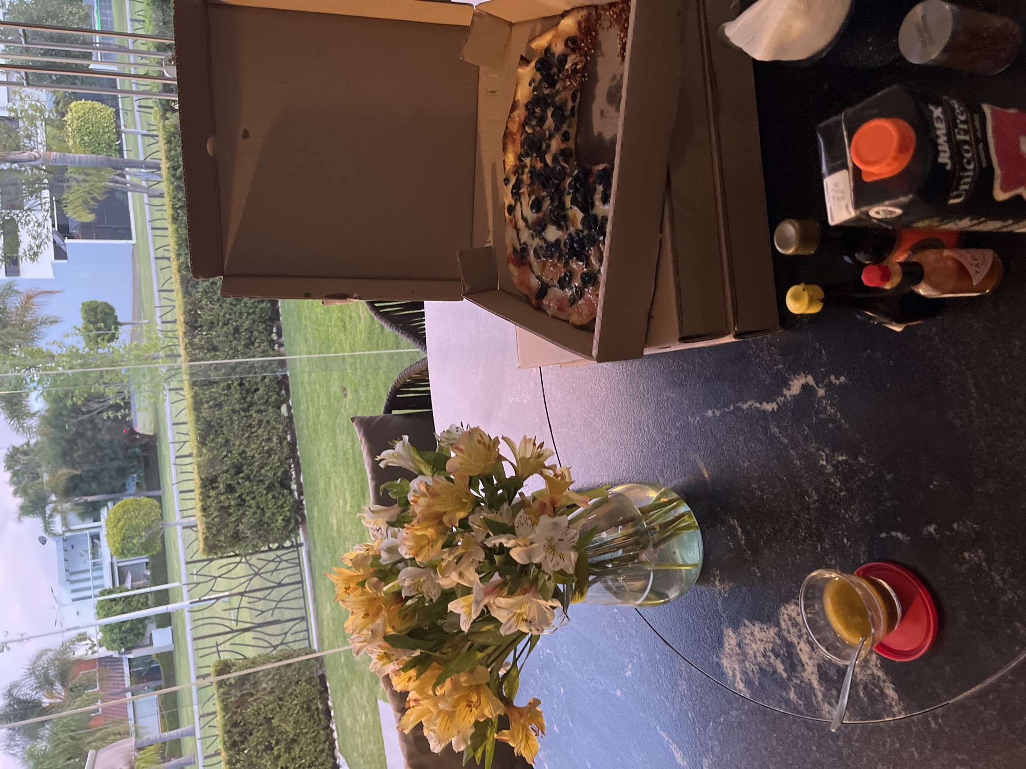 The image shows a table setting with a view of a garden, likely in San Bernardino Tlaxcalancingo, Mexico based on the context. On the table, there is a pizza box with a partially eaten pizza topped with olives, a vase of white and yellow flowers, and an open container with a spoon. Other items on the table include a carton of Jumex juice, a hot sauce bottle, and other small containers with condiments. The scene appears to be set up for a meal outdoors.