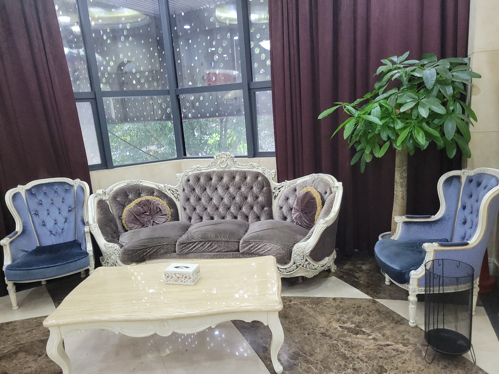 This image captures an opulent indoor seating area, likely a lobby, waiting room, or formal living space, in Liangjiang, China. The central focus is a grand, Victorian-style sofa with a tufted back and armrests, upholstered in a greyish-mauve velvet-like fabric. Its frame is intricately carved with white or silver detailing. Two round throw pillows with gold trim adorn the sofa's corners.
Flanking the sofa are two matching armchairs, also in a Victorian style with tufted backs and carved white/silver frames, upholstered in a striking blue velvet-like fabric. In front of the sofa, a light-colored, possibly marble, coffee table with ornate carved legs displays a small white rectangular tissue box with a subtle floral pattern on its lid.
To the right of the sofa, a tall, leafy green plant, possibly a money tree, stands by the wall. Adjacent to the right armchair, a black metal mesh wastebasket or stand rests on the floor.
The room features large windows with dark frames, covered partially by dark plum or burgundy curtains. The window panes are adorned with numerous reflective, circular elements, creating a distinctive visual effect. Through the windows, lush green foliage and trees are visible, indicating an outdoor view and suggesting it is daytime. The floor is covered with large tiles arranged in a geometric pattern, alternating between dark brown/marble-like squares and lighter, possibly off-white, squares. No people are present in the scene, and no discernible text is visible on any objects. The setting conveys an atmosphere of classic elegance and luxurious comfort.