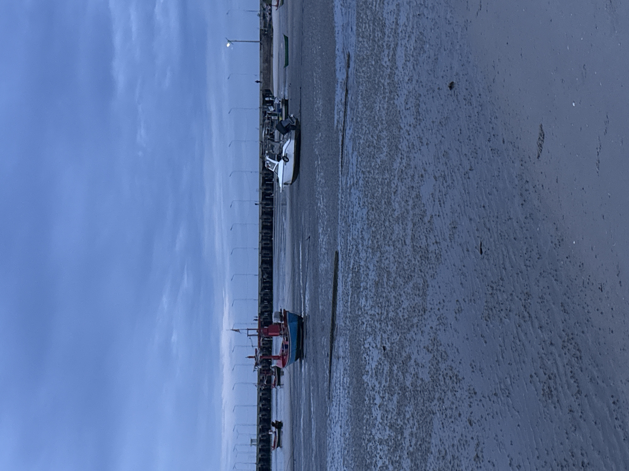 The image depicts a low tide scene at a beach in Hua Hin City Municipality, Thailand. The sky is overcast with dark blue clouds. A long pier extends across the horizon, lined with streetlights. Several boats are scattered on the wet sand and shallow water. In the foreground, the sand is textured with small mounds and ripples. A red and blue fishing boat is prominent in the center, with other smaller boats visible further back, including a white speed boat and a green dinghy. A few indistinct figures can be seen walking on the pier. The overall lighting suggests it is either dawn or dusk.