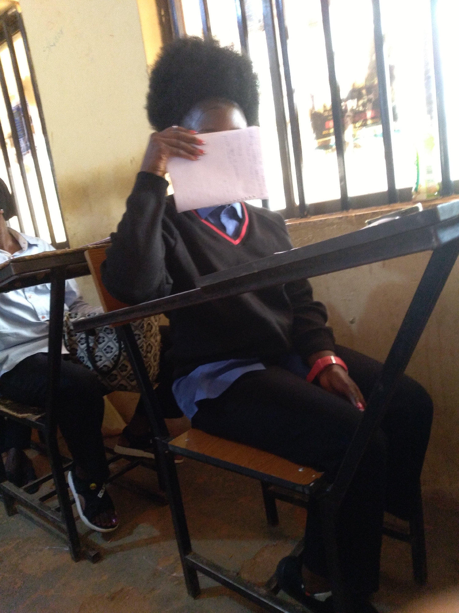 In a classroom in Jos, Nigeria, a person sits at a desk, partially obscured by a piece of paper held in their hand. The individual has dark, curly hair and is wearing a black sweater with a V-neck and a blue undershirt, along with black pants. A red wristband is visible on their left arm. The classroom has metal chairs with wooden seats and backs, and the background features a window with vertical bars, through which bright daylight streams in. Some posters or pictures are visible through the window. The floor is concrete.