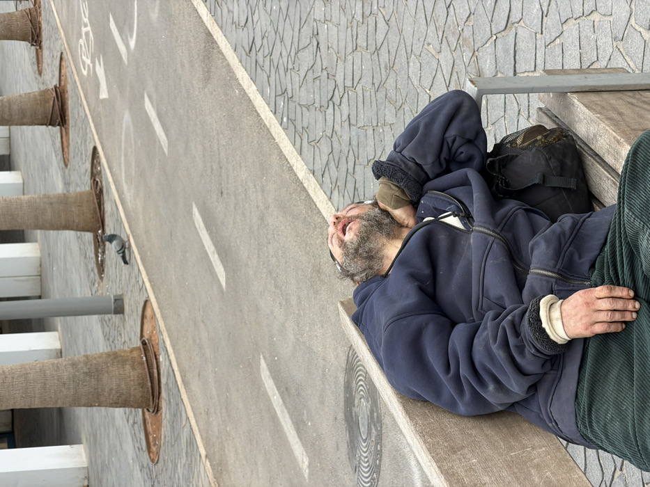 A male individual is depicted sleeping or resting on his back on a light-colored wooden and metal public bench. He has a beard, appears to have disheveled dark hair, and his mouth is open. He is wearing a dark blue zippered hoodie over a lighter colored inner layer, with the cuffs rolled up revealing light fabric, and dark green corduroy pants. His right arm is bent with his hand near his head, while his left arm rests on his lap. A black bag with a mesh pocket is positioned on the bench next to his right hip.
The bench is situated on a paved area with irregularly shaped, light grey cobblestones. To the left of this area is a smooth, dark grey asphalt path marked with white dashed lines and a white bicycle symbol with an arrow, indicating a designated bicycle lane. Beyond the bicycle path, another paved area featuring several evenly spaced palm trees with thick trunks and irrigation rings around their bases is visible. In the background, white, rectangular pillars or columns of a building structure are present. A single pigeon is standing on the paved area next to the bicycle path. A circular manhole cover with concentric ring patterns is visible on the cobblestone pavement near the man's feet.
The scene takes place in a modern public urban space in Tel-Aviv, Israel, specifically resembling the Tel Aviv Port (Namal Tel Aviv) area or a similar renovated promenade with designated pedestrian and bicycle paths, architectural structures, and landscaping.