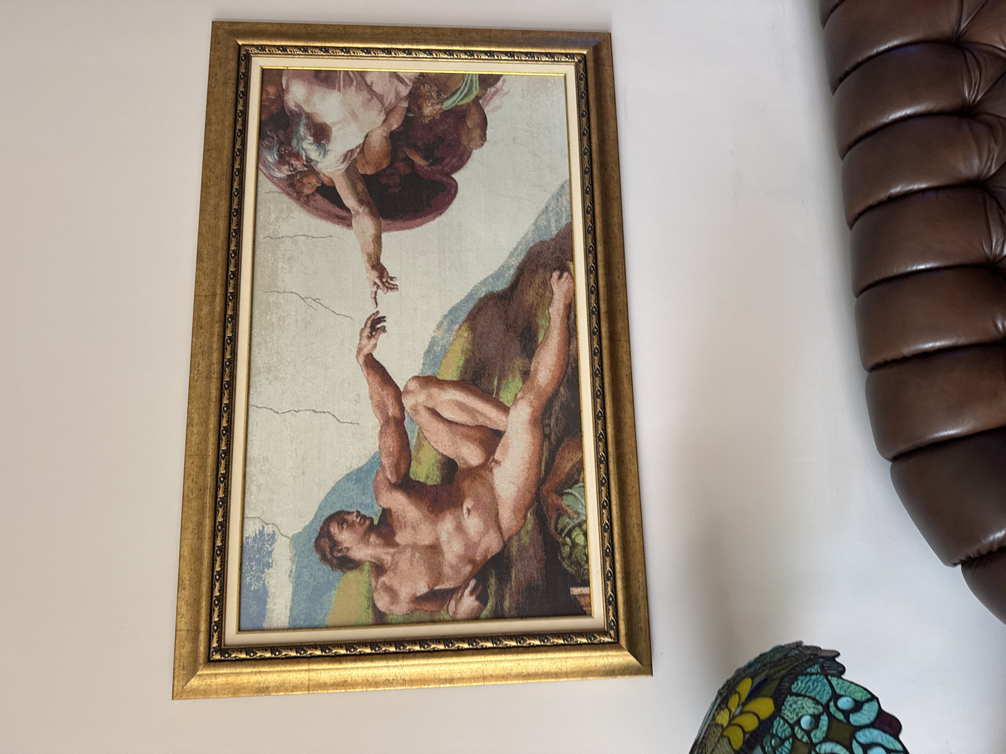 A framed tapestry of Michelangelo's "The Creation of Adam" hangs on a white wall. To the right, part of a brown leather armchair is visible, along with a multi-colored lamp. The location is not discernable from the image, therefore it's impossible to determine if it is in Plovdiv, Bulgaria.