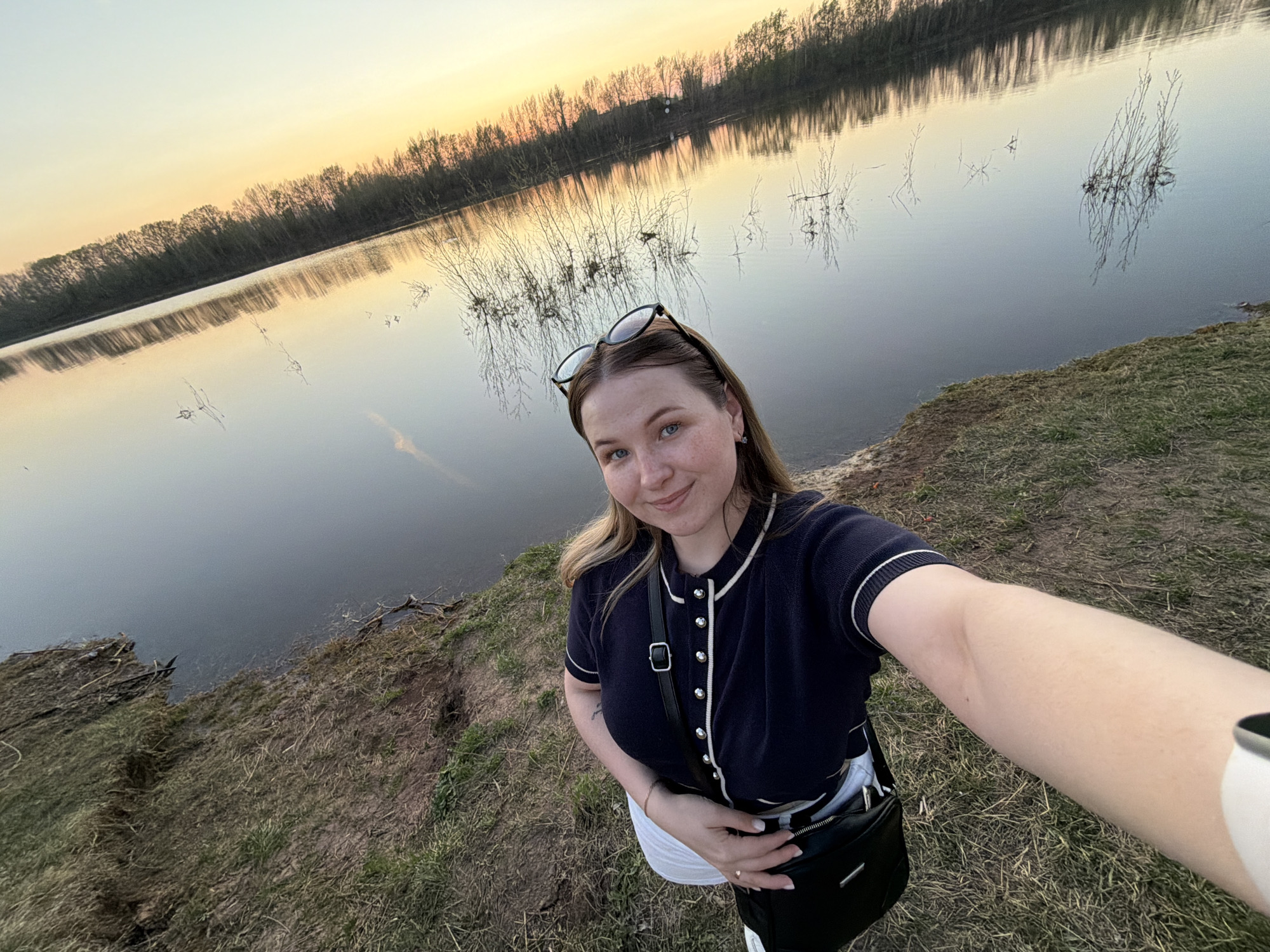 A woman stands on a grassy bank next to a body of water. The location is the city of Chaikovsky, Russia. The sky is a mix of orange and yellow, indicating either sunrise or sunset. The water reflects the sky and the trees on the opposite bank. The woman is taking a selfie. She is wearing a dark blue, short-sleeved cardigan, white pants, and a black shoulder bag. Her sunglasses are perched on top of her head. The bank is covered in dry grass and dirt. Some small branches and bushes are also present near the water's edge.
