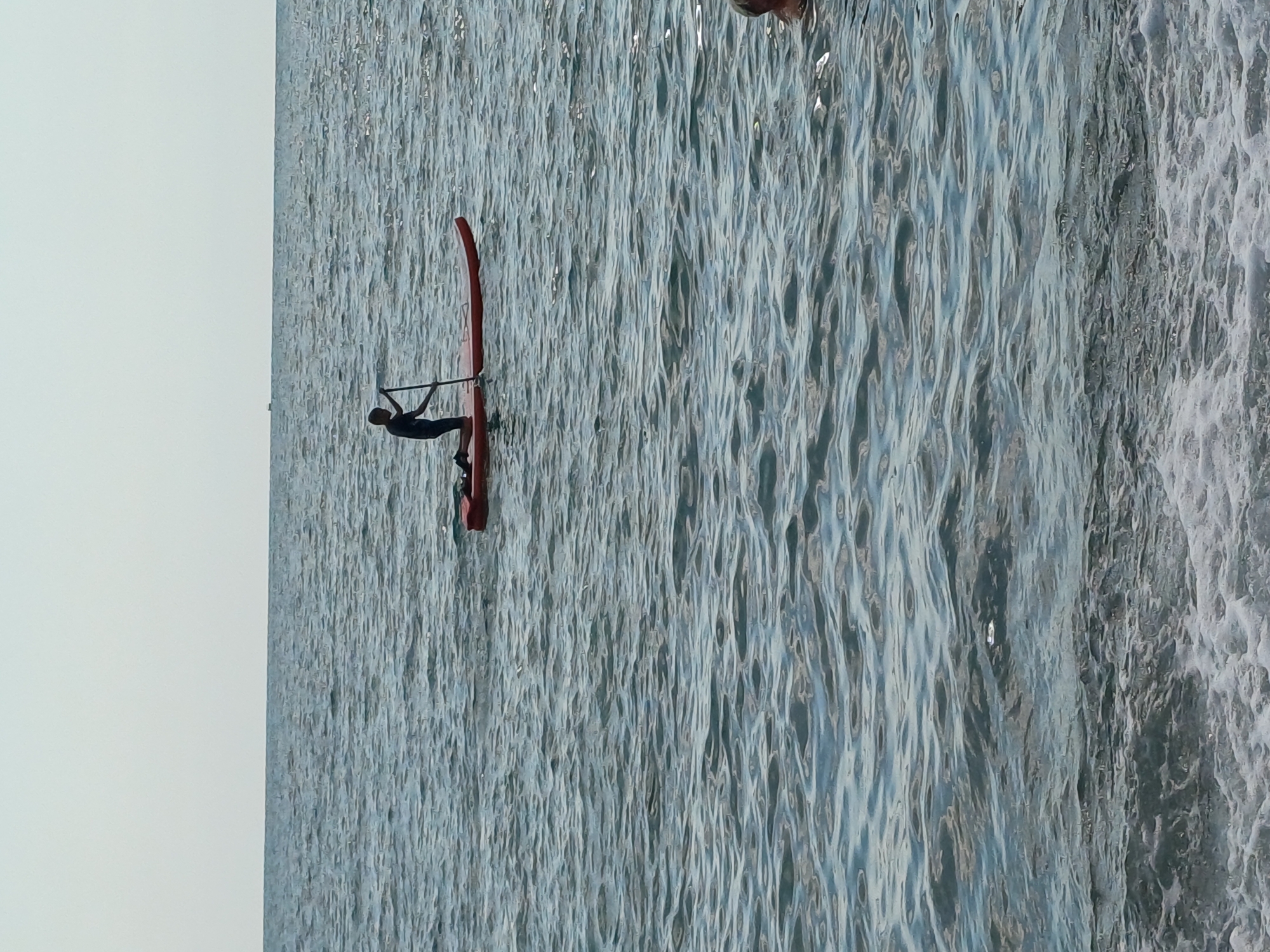 The image shows a person on a red paddleboard on the water. The person is wearing dark clothing and is holding a paddle across their body. The water is choppy with whitecaps, suggesting it may be windy or wavy. The scene appears to be set during the daytime, with bright light reflecting off the water. The background is predominantly water, with a lighter sky on the left side of the image. The person is positioned in the center-left of the frame, actively engaged in paddling. The red of the paddleboard stands out against the blue and grey tones of the water.
