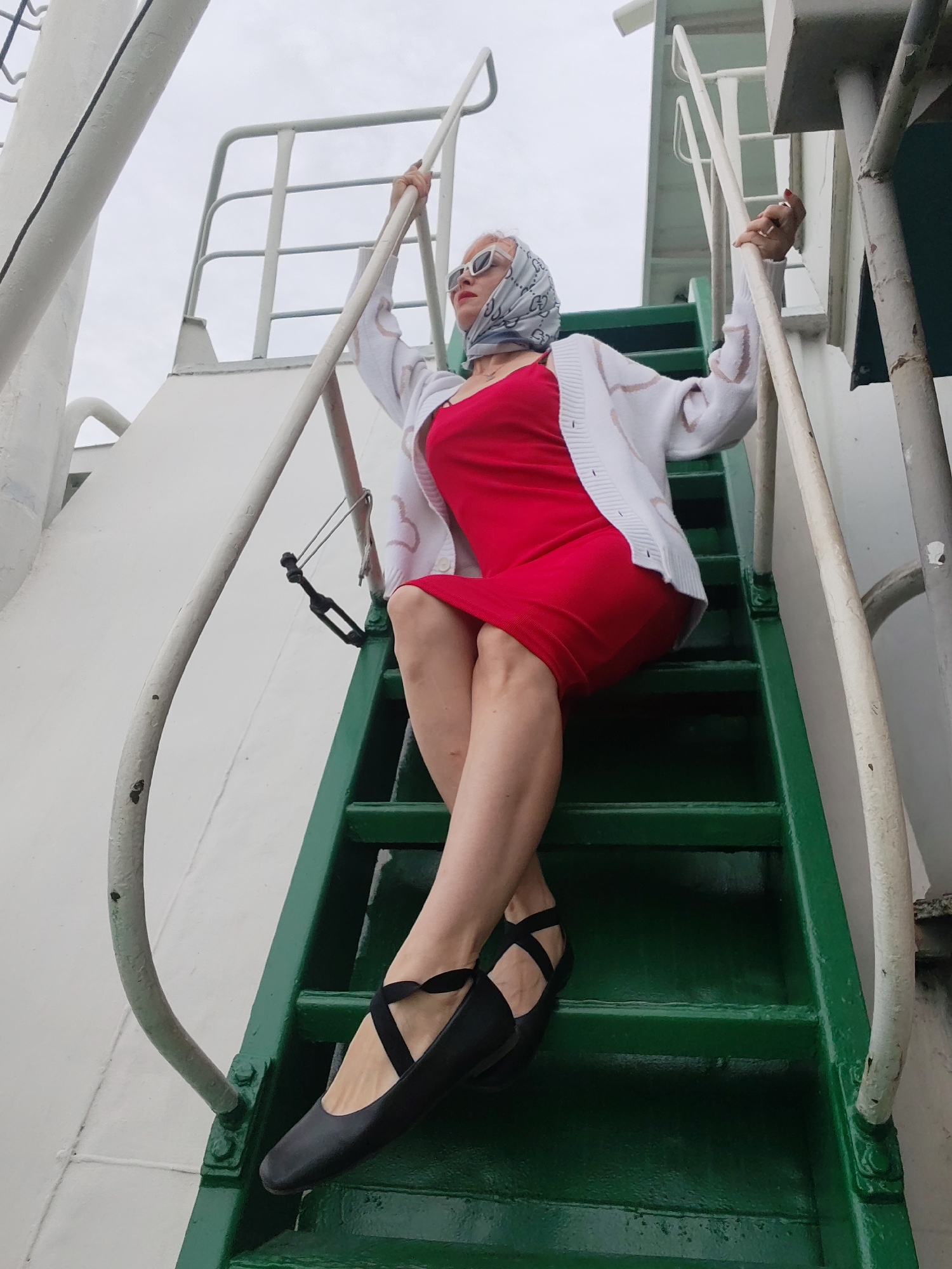 A woman is captured in a full-body shot, from the waist down, posing on a green metal staircase with white handrails. She is wearing a bright red, form-fitting dress and black ballet flats with criss-cross straps. Her legs are crossed at the knees, and one hand is resting on the white railing. She is also wearing a light-colored patterned scarf on her head and white-rimmed sunglasses. The setting appears to be outdoors, possibly on a boat or a structure with industrial elements, suggested by the metal railings and the overcast sky. The overall mood is one of poised confidence and perhaps a hint of glamour. The image was taken in Bereg, Russia.