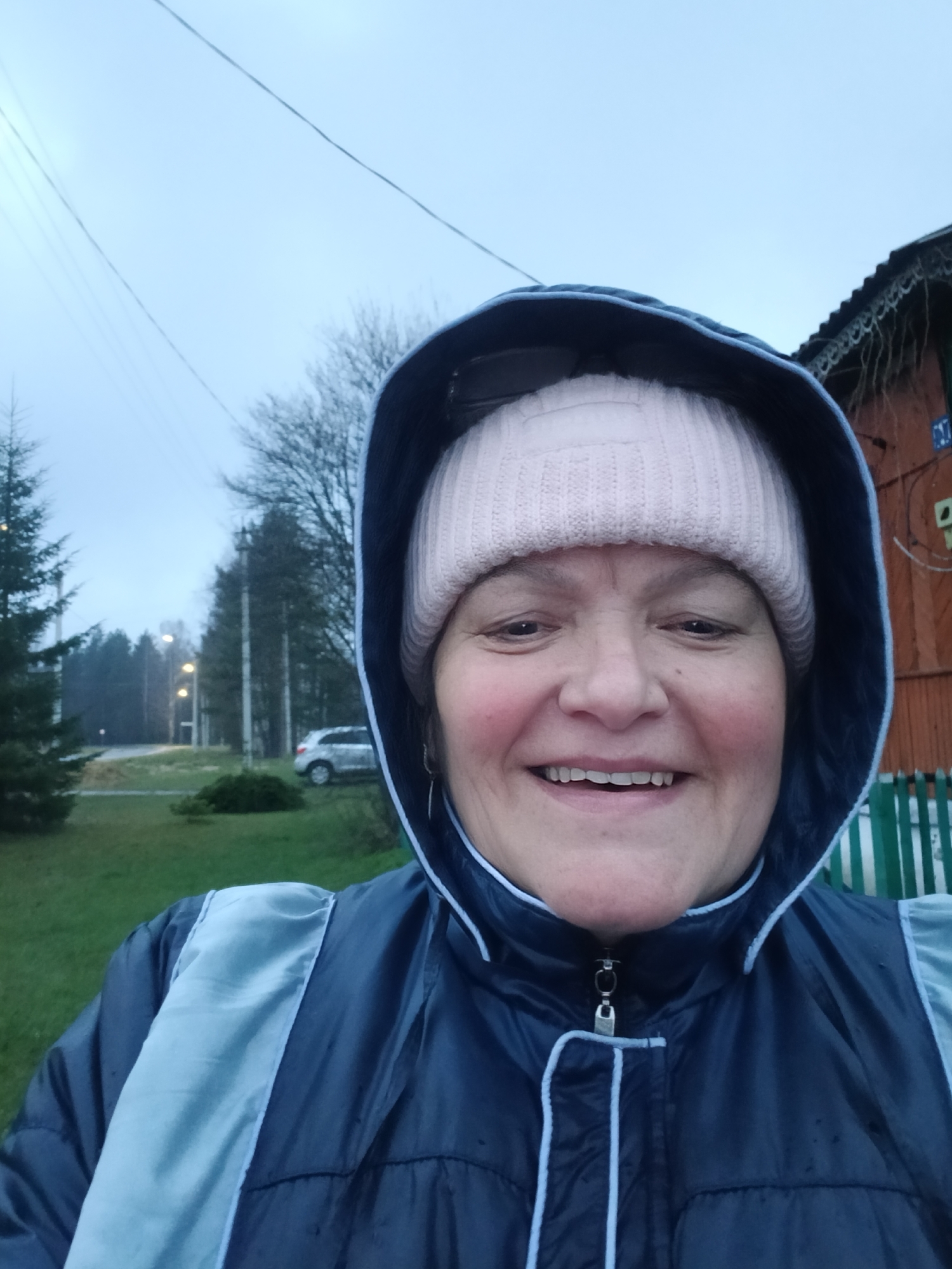 A woman is captured in a cheerful selfie, her face dominating the center of the frame. She smiles broadly at the camera, revealing her teeth, with rosy cheeks complementing her warm expression. She wears a light pink knitted beanie and a dark blue waterproof jacket with the hood drawn up, featuring light blue piping and a vertical light blue panel on the right side of her chest. Small water droplets are visible on her jacket, indicating damp or rainy conditions.
The background on her left shows a green, grassy area with several coniferous trees. Further back, a road is visible with streetlights and power lines stretching across an overcast, light grey sky. A light-colored car is parked on the roadside in the distance. To her right, part of a rustic wooden house is visible, adorned with decorative trim under the eaves and what appear to be icicles or ornate hangings. A green picket fence runs along the front of the house. A small blue sign on the house displays the text "C17".
The overall setting appears to be a rural or semi-rural area in Красный Маяк, Russia, characterized by the simple architecture, natural elements, and sparse infrastructure. The dim, diffused light suggests an overcast day, possibly in the late afternoon or early evening. The woman's bright smile conveys a sense of happiness and resilience despite the potentially dreary weather.