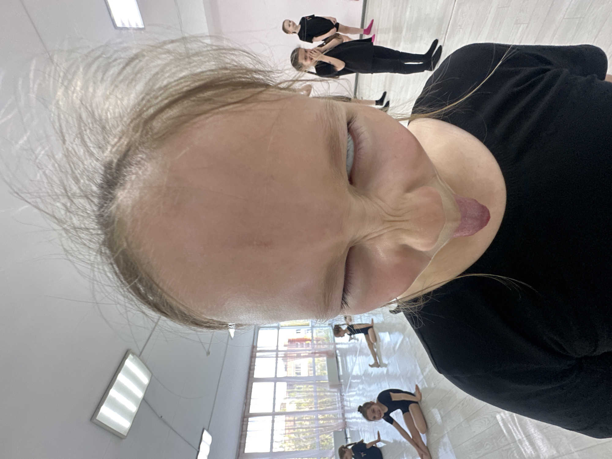 A selfie taken by a young, blonde-haired person in a black shirt in what appears to be a dance studio or gymnasium. The selfie is taken at an awkward angle, focusing closely on their face with their tongue sticking out. Other people in black leotards are stretching in the background. Large windows and rectangular light fixtures are visible in the background. The exact location in Votkinsk, Russia, is indiscernible based on the visual information.