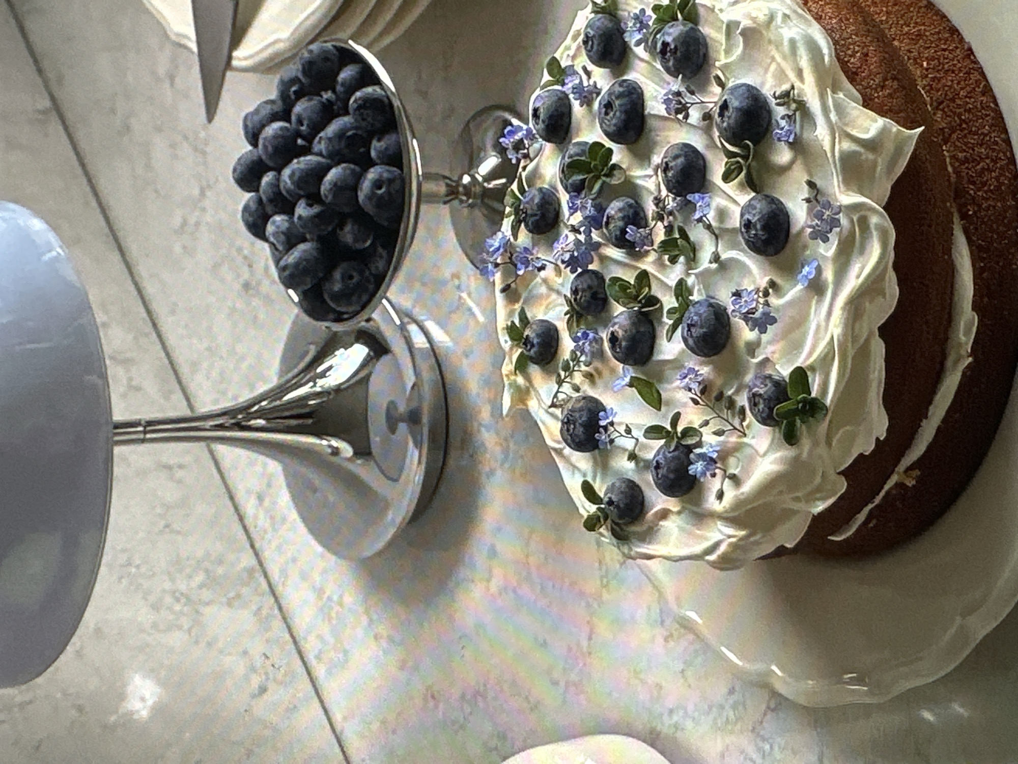 A high-angle shot shows a dessert table set with a layer cake decorated with white frosting, blueberries, and small blue flowers. To the left, a silver pedestal bowl is filled with more blueberries. The scene appears to be indoors with a light-colored surface beneath the cake and bowl. No people are visible. The location in Yerevan, Armenia, is unknown.
