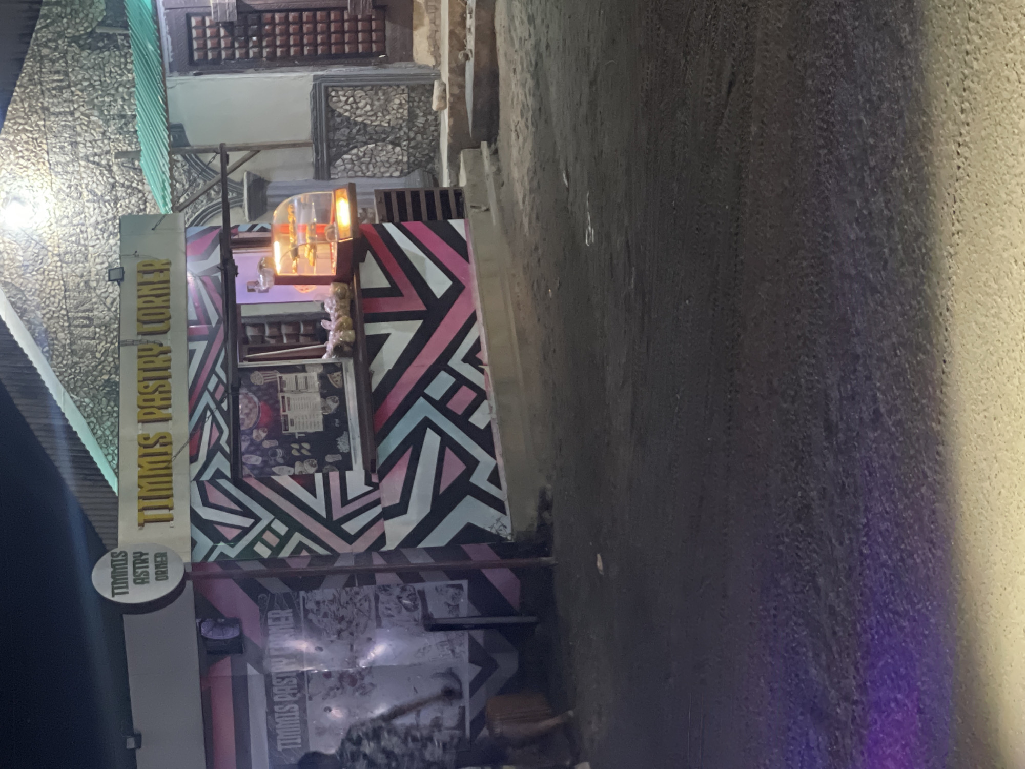 The image depicts "Timms Pastry Corner" in Abraka, Nigeria. The shop is a small, colorful structure with geometric patterns in pink, black, and blue-green. A window displays menu items and baked goods. A sign reading "Timms Pastry Corner" is vertically oriented on the shop's exterior. There is a poorly lit street adjacent to the shop. People are present near the shop. There are buildings around the pastry shop.