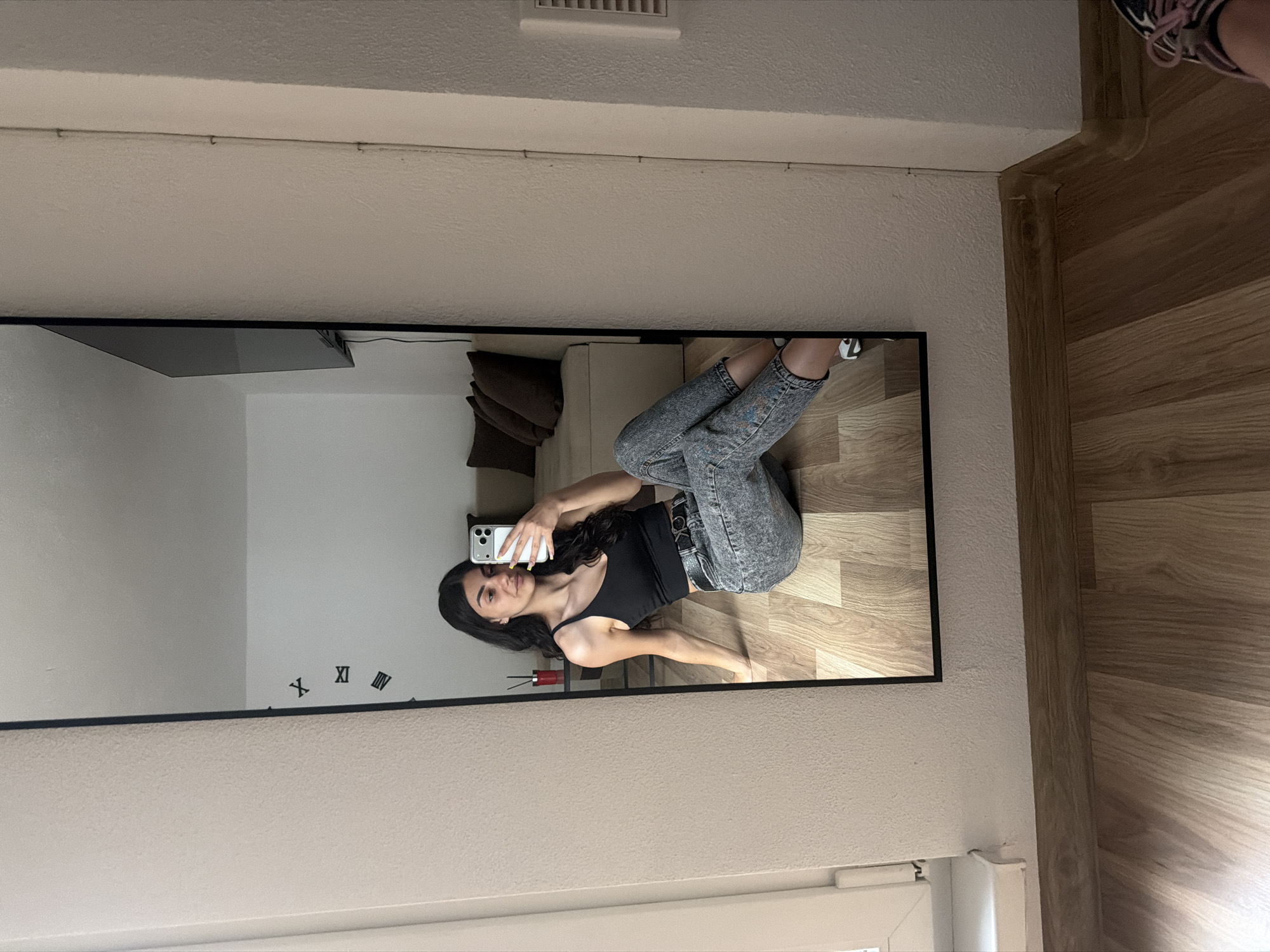 A young woman with long, dark hair is captured in a mirror selfie within an indoor setting in Gabrovo, Bulgaria. She is wearing a black tank top and light-colored acid-wash jeans, secured by a belt featuring the letters "CK" (Calvin Klein) on the buckle. She holds a smartphone, with its multiple camera lenses visible in the reflection, up to take the picture, maintaining a neutral expression.
The large rectangular mirror, framed in black, reflects a room with light-colored walls and wooden flooring. Visible within the mirror's reflection are a beige sofa adorned with brown throw pillows and a red cylindrical decorative item. Part of a wall clock, showing Roman numerals "X" and "XI", is also reflected. Outside the mirror, sections of the room's ceiling with a white air vent, a textured light wall, and wooden flooring are visible, along with a partial view of a white window frame at the bottom right. The image captures a casual, everyday moment in a home environment.