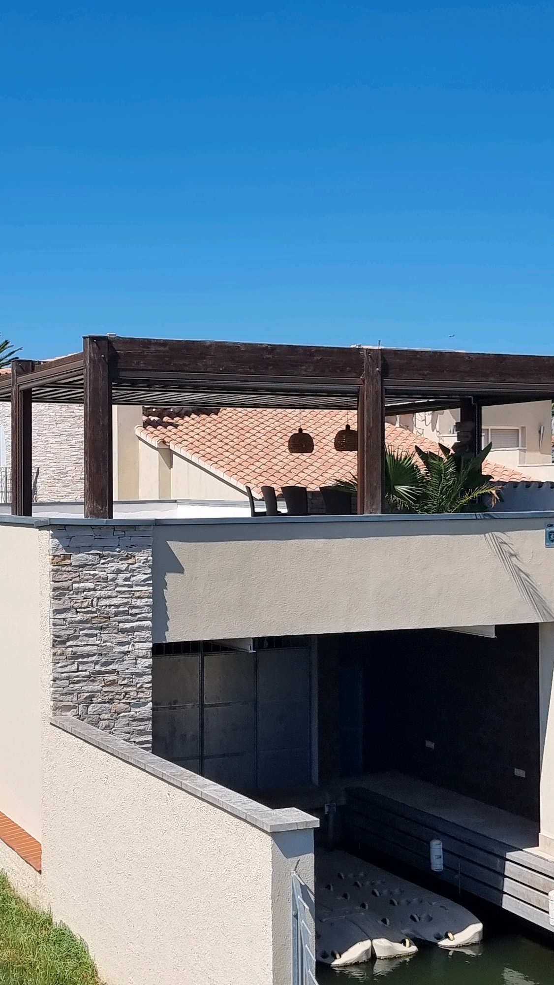 The image shows a modern waterfront property in Castelló d'Empúries, Spain. The foreground features a cream-colored building with a textured finish, leading to a water channel where two dark, boat-like structures are moored. Above this, a white wall with a stone-clad section creates a terrace area. Dominating the upper portion of the image is a dark wooden pergola structure extending over a tiled roof of a building. Beneath the pergola, patio furniture and decorative pendant lights are visible. The scene is set against a clear blue sky.