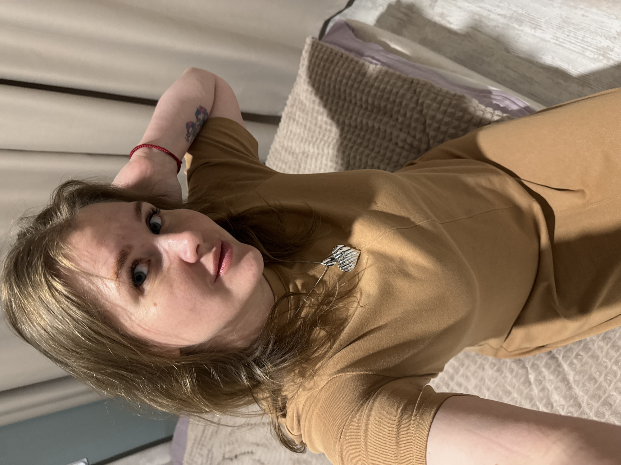 A young woman with light brown hair and fair skin is depicted in a close-up, low-angle selfie, seemingly taken while she is lying down. She looks directly into the camera with a composed, neutral expression. Her hair frames her face, and her light-colored eyes are prominent. She wears a high-necked, long-sleeved top and what appear to be matching pants, both in a solid camel or tan color. A silver-toned, abstract heart-shaped pendant or brooch is pinned to her top near her collarbone. On her left wrist, a delicate red beaded bracelet is visible, and a small, colorful tattoo featuring floral or abstract designs is present on her left upper arm. Her left arm is bent, with her hand resting behind her head in a relaxed pose. She is positioned on a bed or sofa, indicated by a textured, light-colored pillow or cushion and light-colored bedding with a subtle pattern in the background. The scene is illuminated by soft, indirect lighting, casting gentle shadows and suggesting an indoor setting, possibly during the daytime. No visible text is present in the image. The implied location context is Gornyi (Dvurechenskoe S/P), Russia, though no specific visual cues confirm this within the photo.