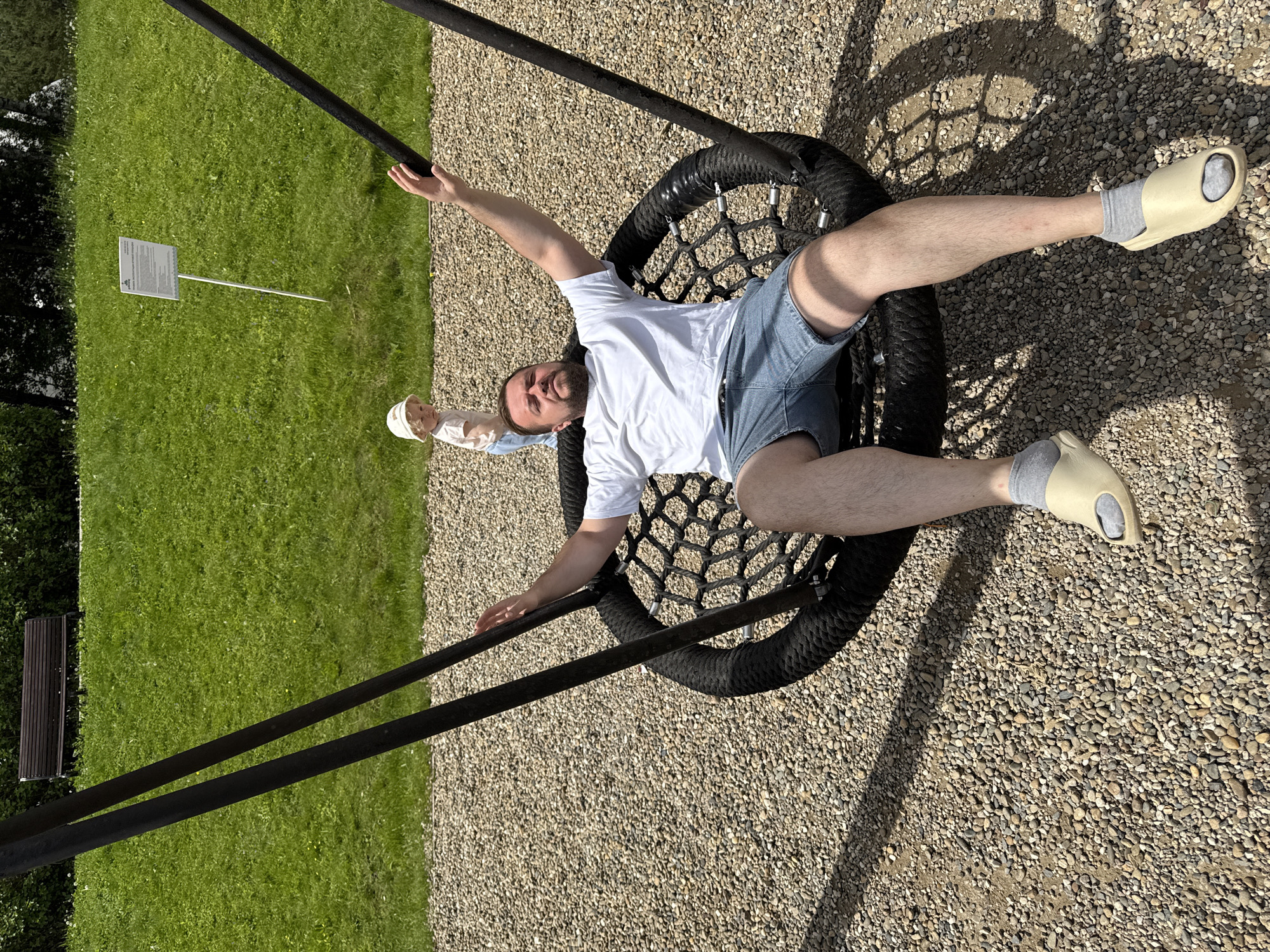 A full-length, overhead shot captures a man lying face-up on a large, round nest swing in an outdoor setting in Dobrograd, Russia. He is positioned centrally within the black woven swing, with his arms and legs spread outwards, giving a relaxed or playful appearance. The man wears a white short-sleeved t-shirt, light-wash denim shorts, and cream-colored slip-on sandals with grey socks. He has a beard and is looking towards the camera.
A young child, partially visible in the upper left corner near the man's head, stands upright and wears a white hat. The ground beneath the swing is covered in light-colored gravel, contrasting with the vibrant green grass and bushes in the background. Strong sunlight creates distinct shadows of the swing structure and the man on the gravel, indicating a clear, sunny day.
In the grass to the left, a small white sign on a thin stake is visible, though its text is illegible. Further back on the grass, a dark wooden slatted bench can be seen. The swing's support bars, also black, extend upwards and out of frame. The scene conveys a leisurely atmosphere in a park or playground environment.