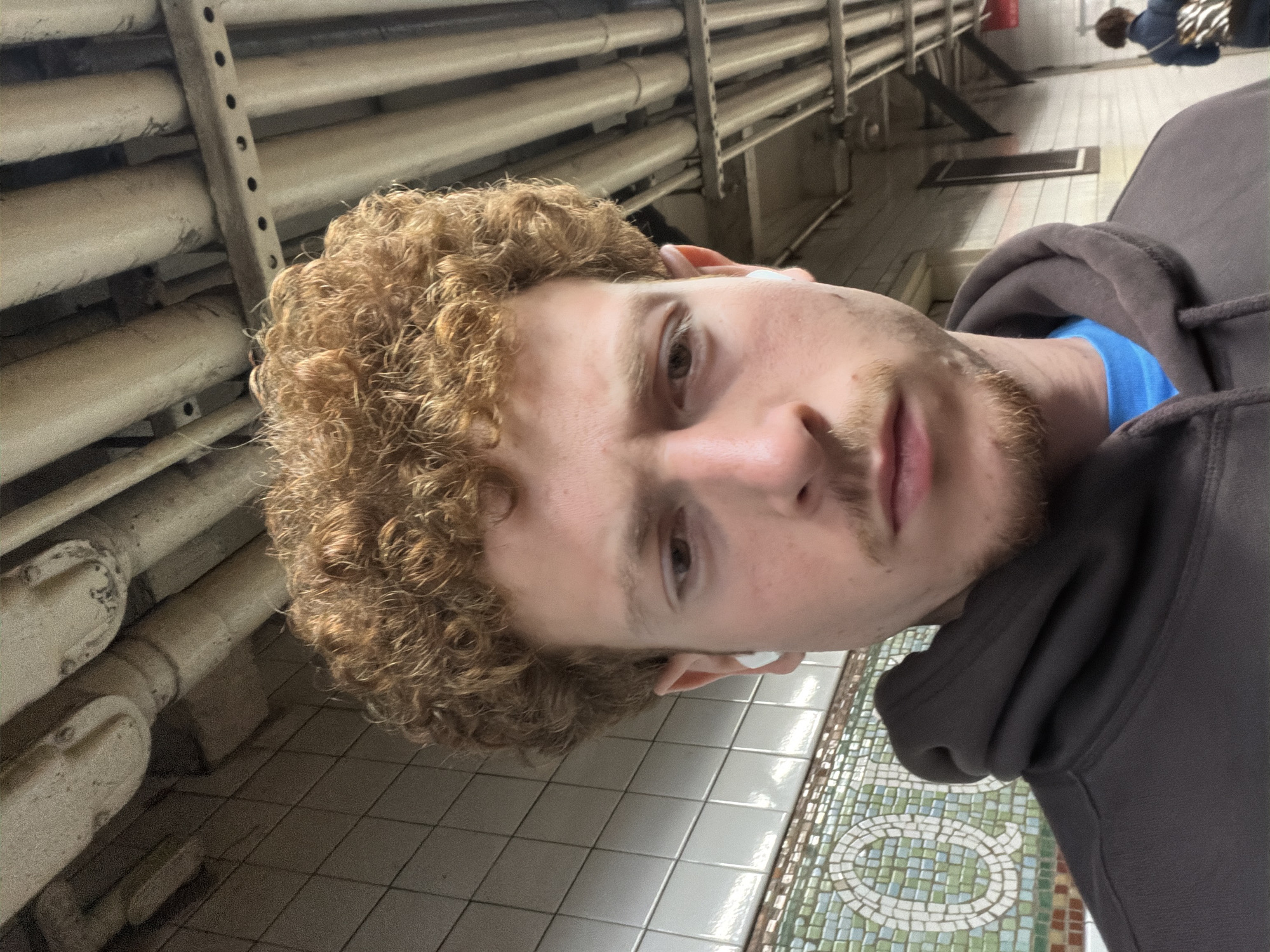 The photograph shows a person with curly red hair, wearing a grey hoodie, standing in a New York City subway station. The person has a white object in their right ear, possibly an earbud. The background shows a tiled wall and numerous pipes running horizontally. The floor is tiled with a mosaic pattern. Other people are visible in the background but are out of focus.