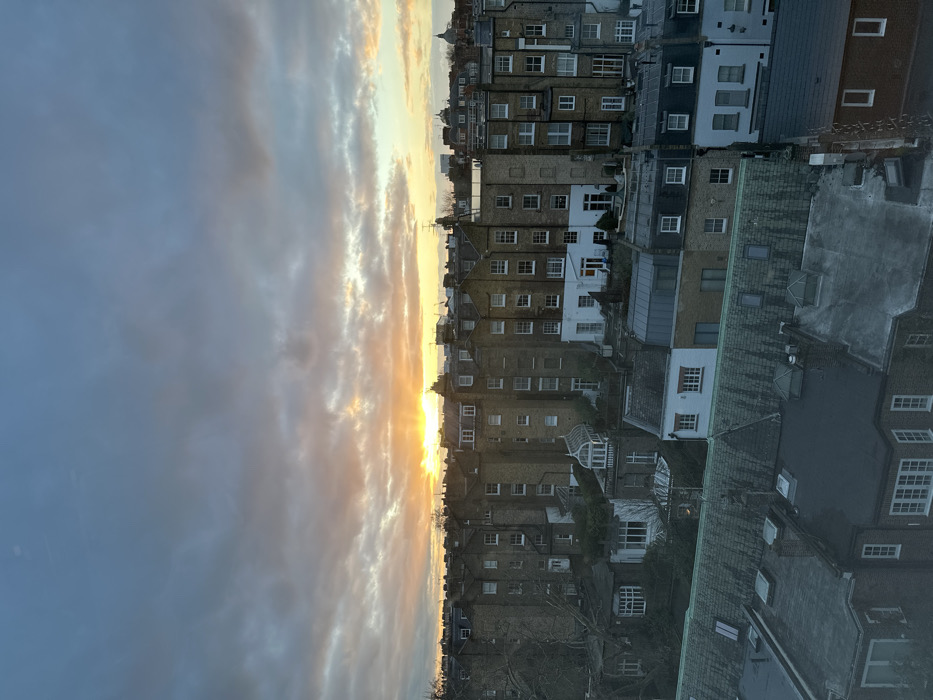 A row of tightly packed buildings stretches along the horizon under a dynamic sky. The buildings vary in height and color, with brick and white facades, some displaying rows of windows. The sky transitions from a dark, cloudy blue to a bright, orange-tinged glow where the sun is setting, casting long shadows. The angle of the photo gives a sense of height, as if looking out from a window over the city.