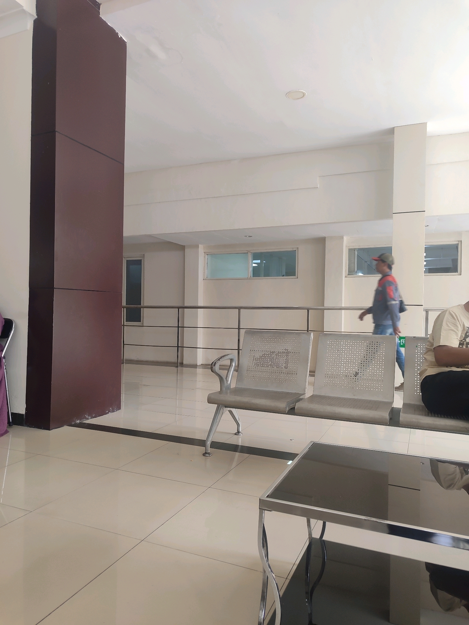 The image depicts an indoor waiting area, likely in a public building. A large, dark brown pillar stands prominently on the left side of the frame. The floor is tiled with large, light-colored square tiles, with a black border line running along the base of the pillar. In the mid-ground, several rows of metallic, perforated chairs are arranged. A person wearing a blue shirt, jeans, and a cap is walking away from the camera towards the right, across the tiled floor and behind the chairs. To the far right, a person is seated, facing away from the camera, wearing a light-colored t-shirt. In the background, a railing separates the waiting area from a hallway or corridor with windows. The walls are painted a neutral, off-white color, and the ceiling is also white with recessed lighting. The overall impression is one of a clean, modern, and functional space. Without further context or specific identifying markers within the image, it is impossible to definitively pinpoint this location to the city of Surabaya, Indonesia.