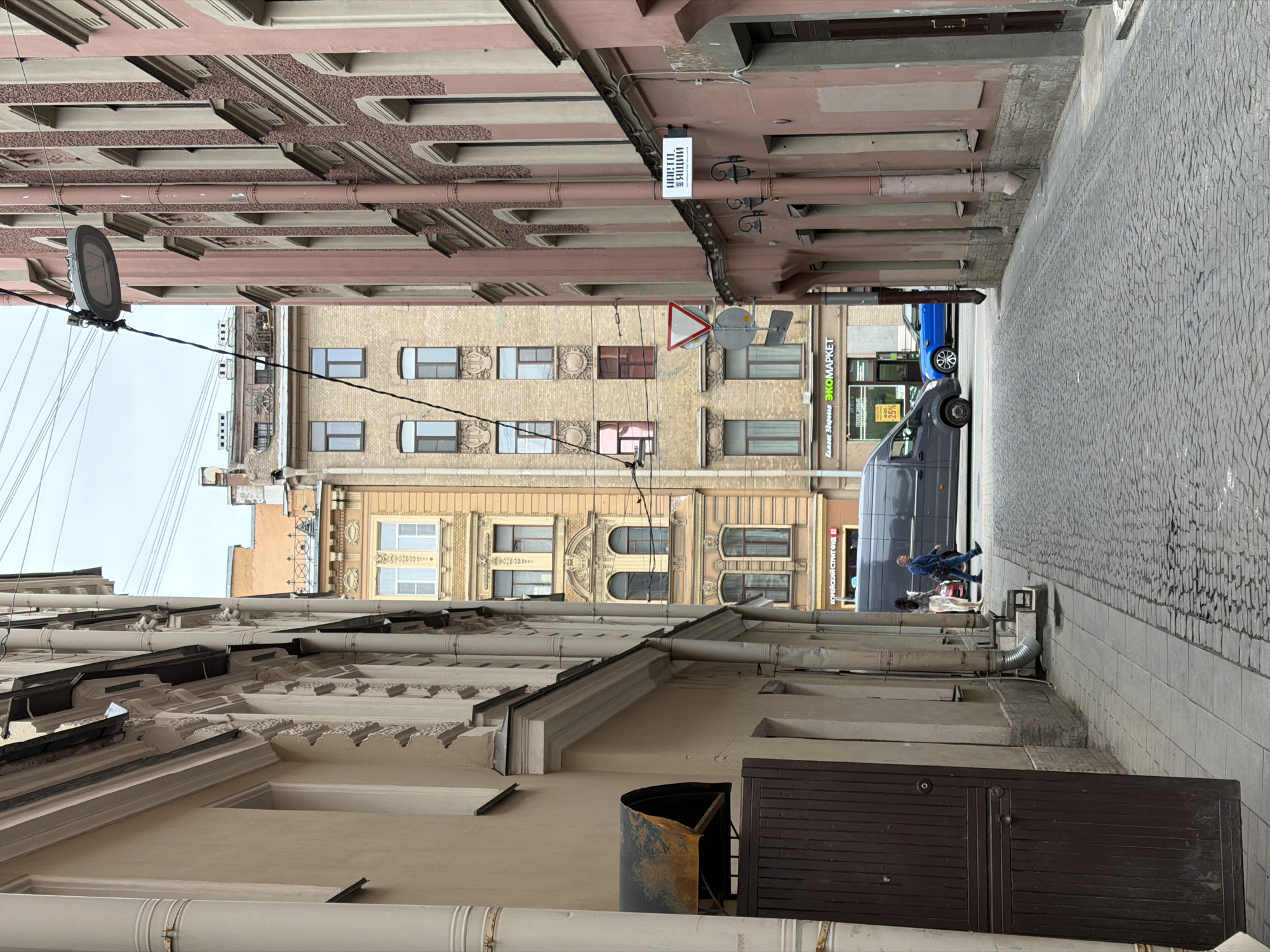 The image depicts a narrow street scene in Saint Petersburg, Russia. The street is paved with gray cobblestones. The view is framed by buildings on either side. The buildings are several stories high and have varying architectural styles. The building on the left has light-colored facade with decorative elements, while the building on the right is painted a pinkish beige with rows of pipes. A grey van and a blue car are parked along the street. A person is walking near the van. Overhead, a light fixture is attached to a wire spanning the street, as well as street signs.