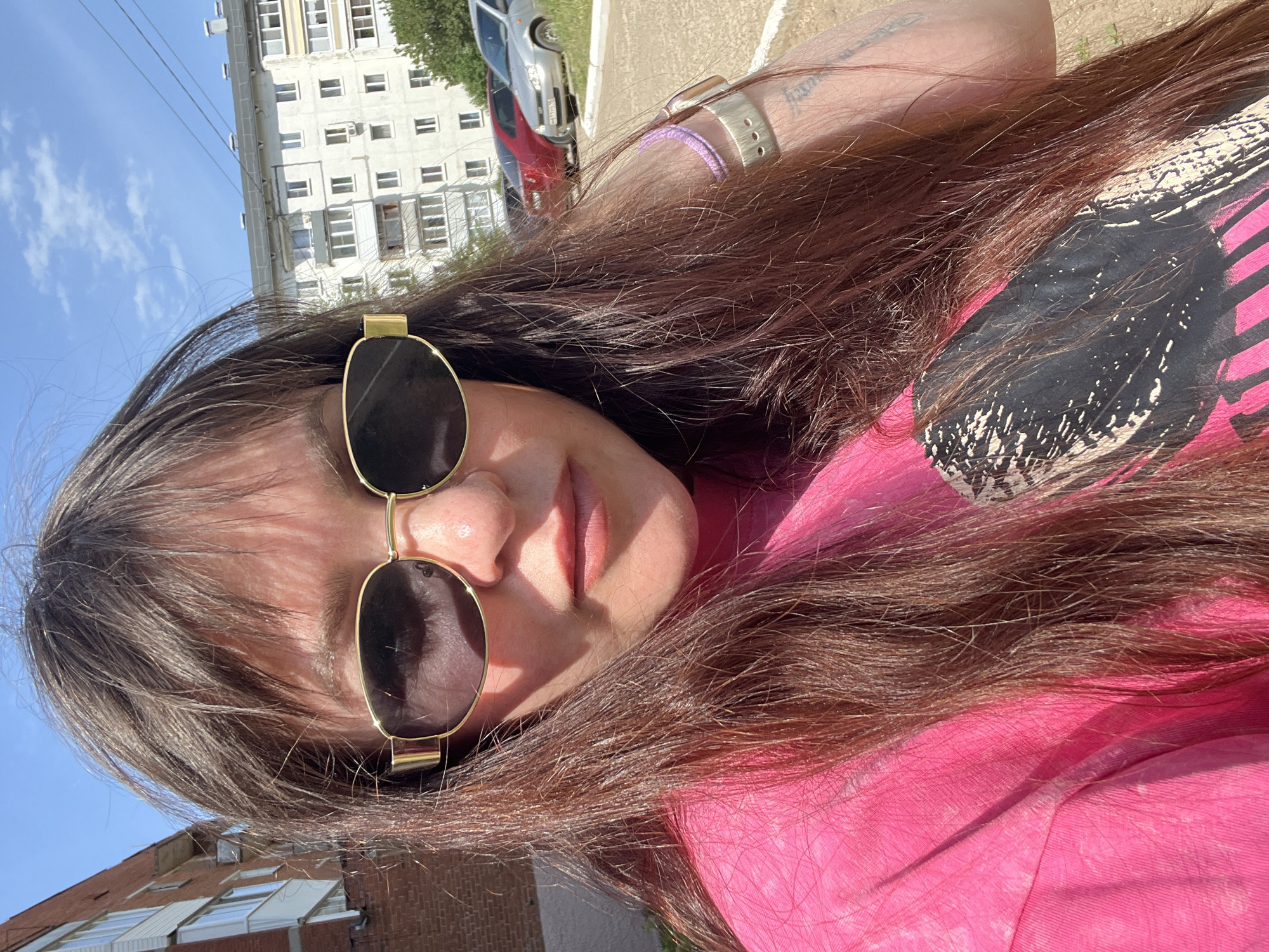 A woman with long brown hair and sunglasses is looking at the camera, wearing a pink shirt with a black and white design. She is outside in Noviy, Russia, as evidenced by the building in the background. There are cars parked in the distance. The photo is taken during the day in sunlight. The woman has a watch on her left wrist.
