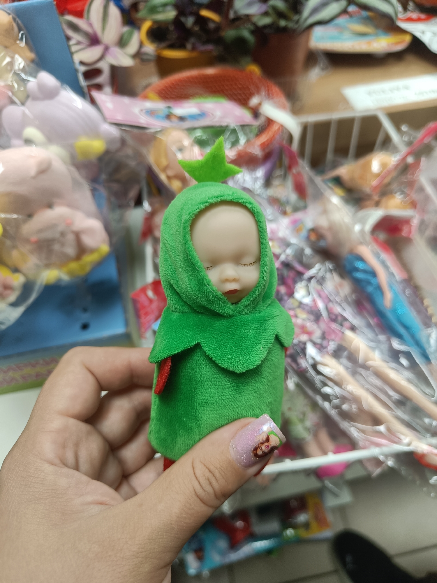 A person's hand holds a small doll dressed in a green fuzzy outfit with a hood. The doll has a pale face and closed eyes, with red lips. A red detail is visible on the doll's outfit. In the background, shelves are filled with various toys and items, including stuffed animals, dolls in packaging, and other colorful objects. The setting appears to be a retail environment, possibly a toy store or a market stall in Krasnodar, Russia. The lighting suggests it could be daytime. The person's fingernail has a small decal of a person's face on it.