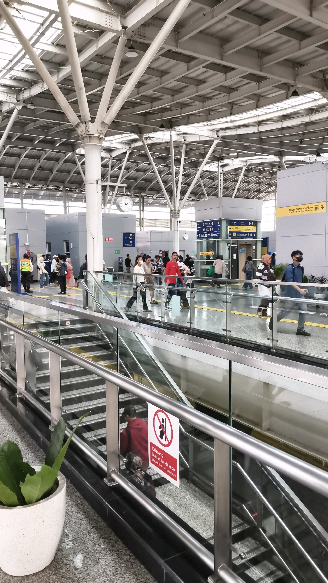 The image depicts the interior of a modern train station in East Jakarta, Indonesia. The scene is a well-lit, spacious concourse with a high, exposed ceiling supported by a complex network of white metal beams. Natural light filters in from above.
In the foreground, a metal and glass railing guides the viewer's eye toward a set of stairs and an escalator, both made of metal and dark material. To the left, a large white planter holds a lush green plant. A prominent red and white sign with a pictogram and text in Indonesian and English ("Dilarang bersandar di kaca / Do not lean on the glass") is attached to the railing.
The midground shows a busy platform area where several people are walking. They are dressed in casual clothing, and some are wearing face masks. There are signs indicating directions and platform numbers, such as "Kijar Commuter Line" and "4-8". People are seen walking in various directions, some appearing to be commuters.
The background reveals more of the station's architecture, including pillars, walls, and further signage. The overall impression is one of an active transportation hub.