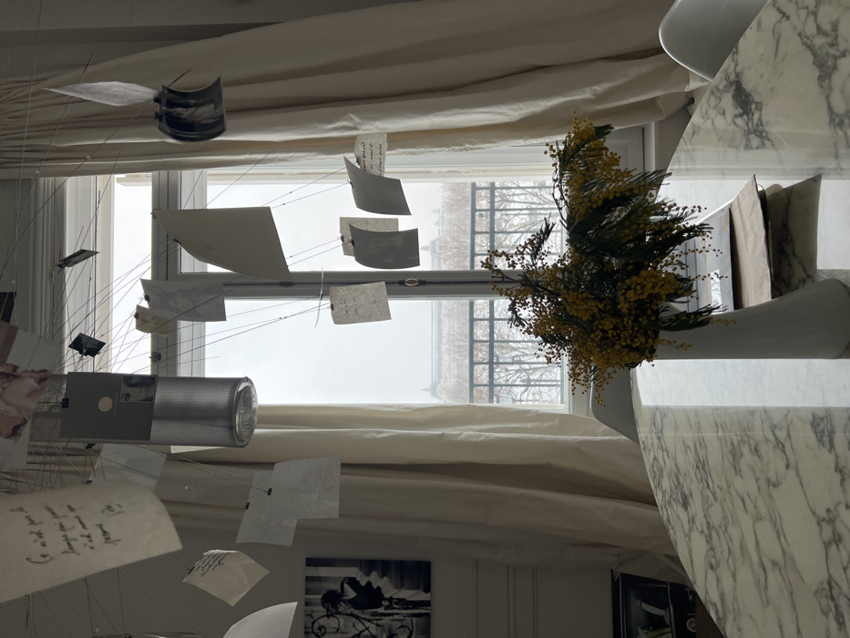 The image displays an interior room, likely an apartment, offering a view of a snow-covered urban landscape in Paris, France. In the foreground, a reflective white marble table with grey veining is centered. On this table, a white, conical vase holds a dense arrangement of yellow mimosa flowers and green foliage. A portion of a white chair is visible on the right side of the table.
Above the table, an elaborate mobile or art installation is suspended, consisting of multiple small pieces of paper, photographs, and grey rectangular items attached by thin wires or strings. Some papers bear handwritten text, with one piece clearly showing "Ce n'est pas le temps qui passe" in French. A metallic cylindrical object is integrated into this overhead structure. The background is dominated by a large window framed by cream-colored drapes. On the left wall, a black-and-white print depicting a woman next to classical architectural columns is visible.
Beyond the window, a black metal balcony railing is present. The exterior scene reveals a snow-covered environment with bare trees and a street or park. In the mid-distance, the distinctive large dome of the Hôtel des Invalides is clearly visible, positioning the interior scene within a building overlooking the Esplanade des Invalides, likely in the 7th arrondissement of Paris. The sky is overcast and bright. No individuals are present in the image.