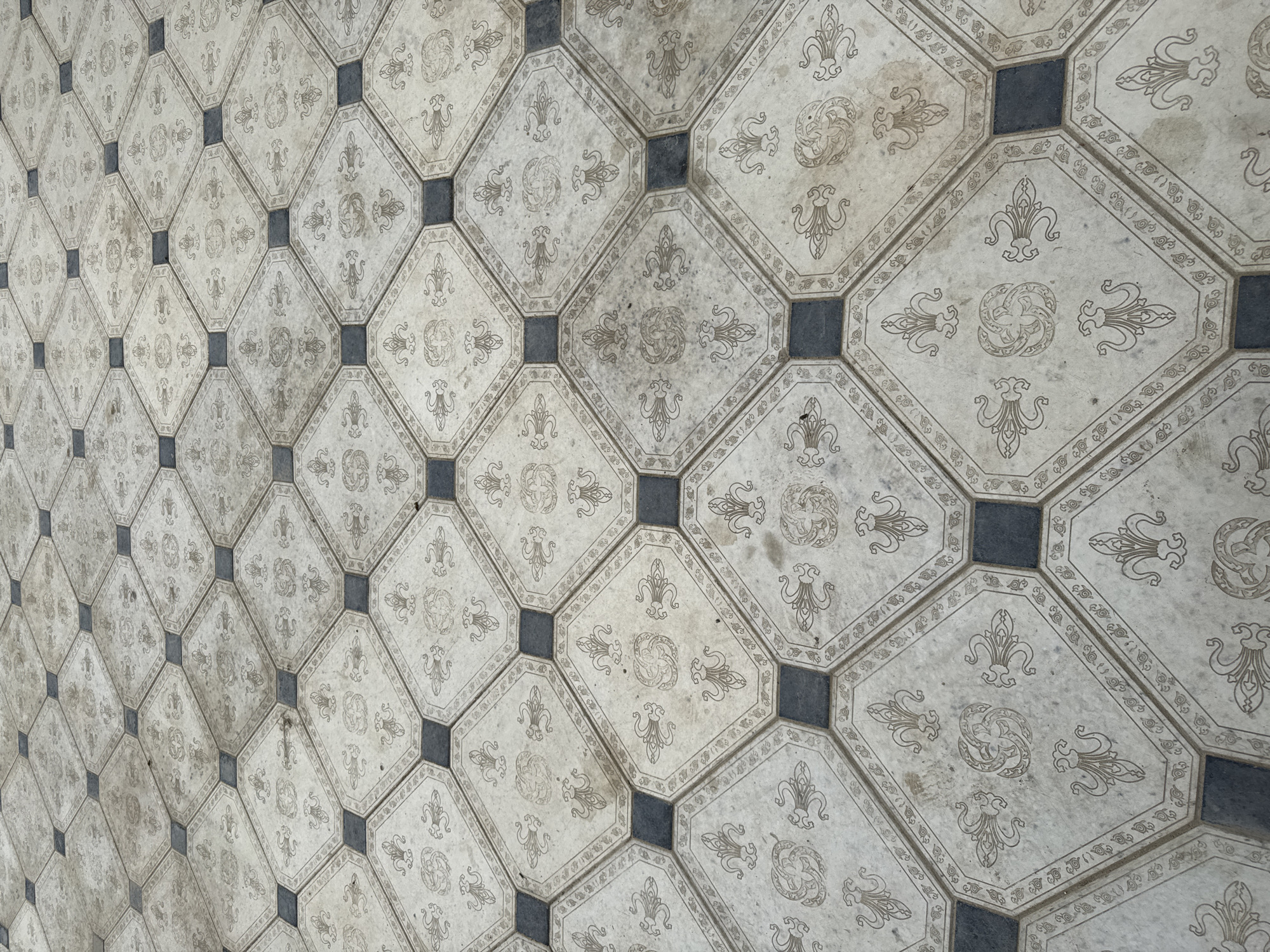 The image shows a close-up, eye-level view of an outdoor tiled floor. The tiles are arranged in a repeating pattern of octagons with decorative designs and small square inserts in dark gray. The octagonal tiles are light beige with intricate, repeated patterns in a darker brown or gray, including what appear to be stylized floral motifs and circular designs. The overall impression is of an aged or weathered surface, with some areas of discoloration and a visible layer of dust or dirt. The perspective is from above, looking down at an angle, and the floor extends into the distance, creating a sense of depth. There are no people or specific landmarks visible. The image was taken in the city of Beshtal, Uzbekistan.