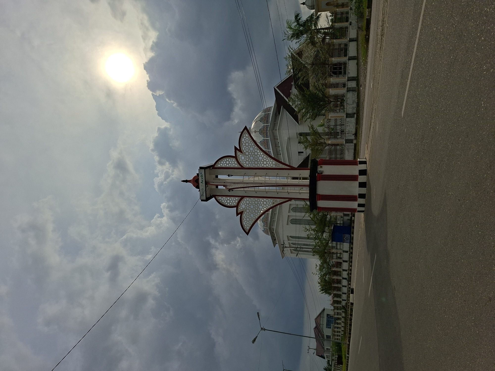 A roadside view in Meunasahaluedrienlhoksukon Barat, Indonesia. The camera faces a tall, ornate structure with a red and white base on the right side of the street. A mosque with a rounded dome stands behind the structure. A road stretches away from the camera on the right, with buildings and trees lining it. Utility poles are also visible along the road. The sky is filled with clouds, and the sun shines brightly in the upper left corner of the image.