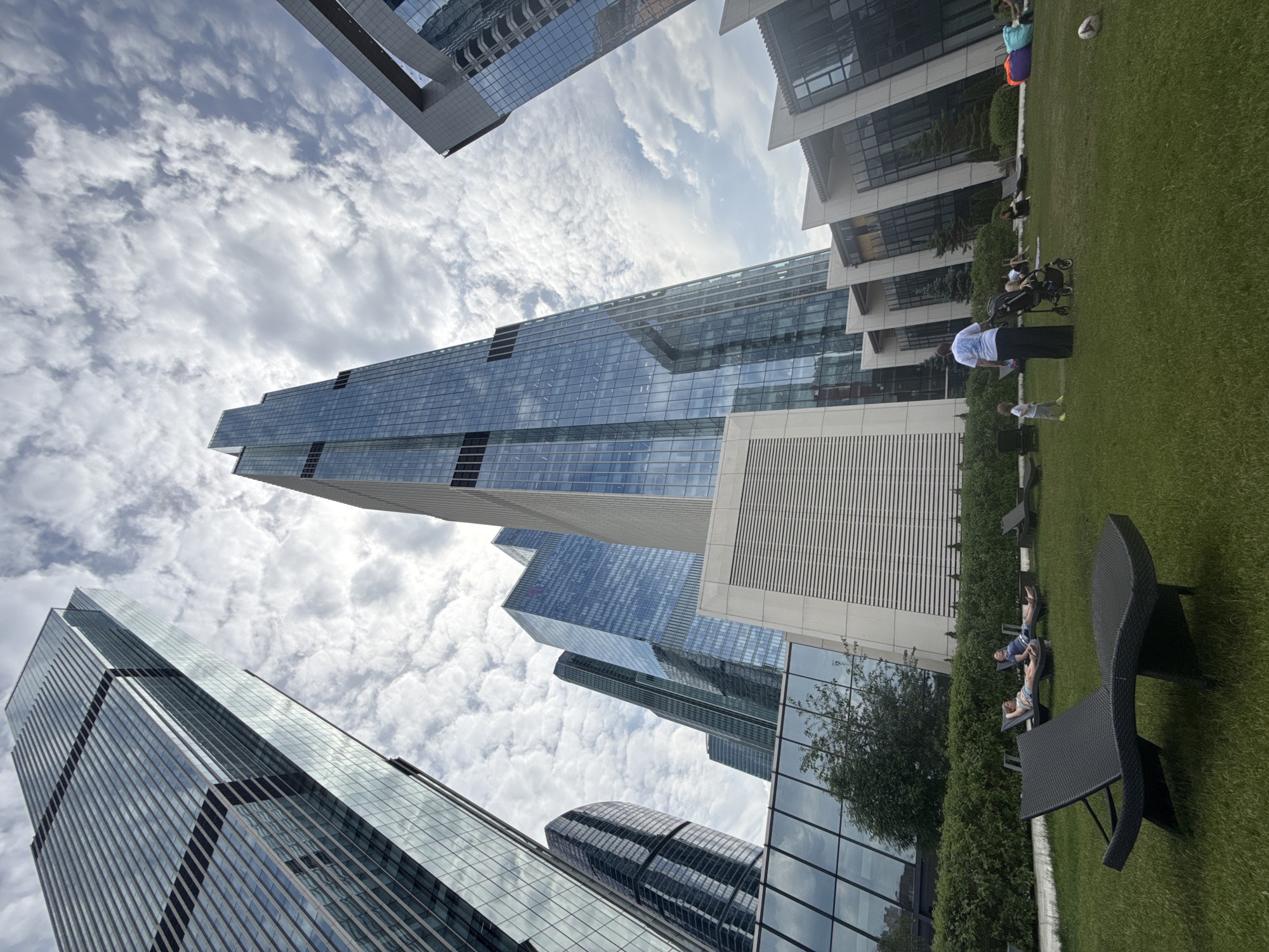 This is a low-angle, wide shot of a modern, upscale outdoor area with several people enjoying a cloudy day. In the background, towering glass skyscrapers dominate the skyline, reflecting the sky and clouds. The foreground features a meticulously manicured green lawn with several black, modern-style lounge chairs. Some people are seen relaxing on the lounge chairs.
In the middle ground, a woman in a white t-shirt and dark pants stands next to a stroller, facing away from the camera. A small child in a light-colored t-shirt and shorts is standing to her left. Other people are scattered across the lawn, some sitting on the grass, others on lounge chairs. There is a rugby ball on the grass in the far right of the image.
The overall impression is one of urban leisure and modern architecture. The location is most likely Moscow City, Russia, a business district known for its contemporary skyscrapers and surrounding amenities.