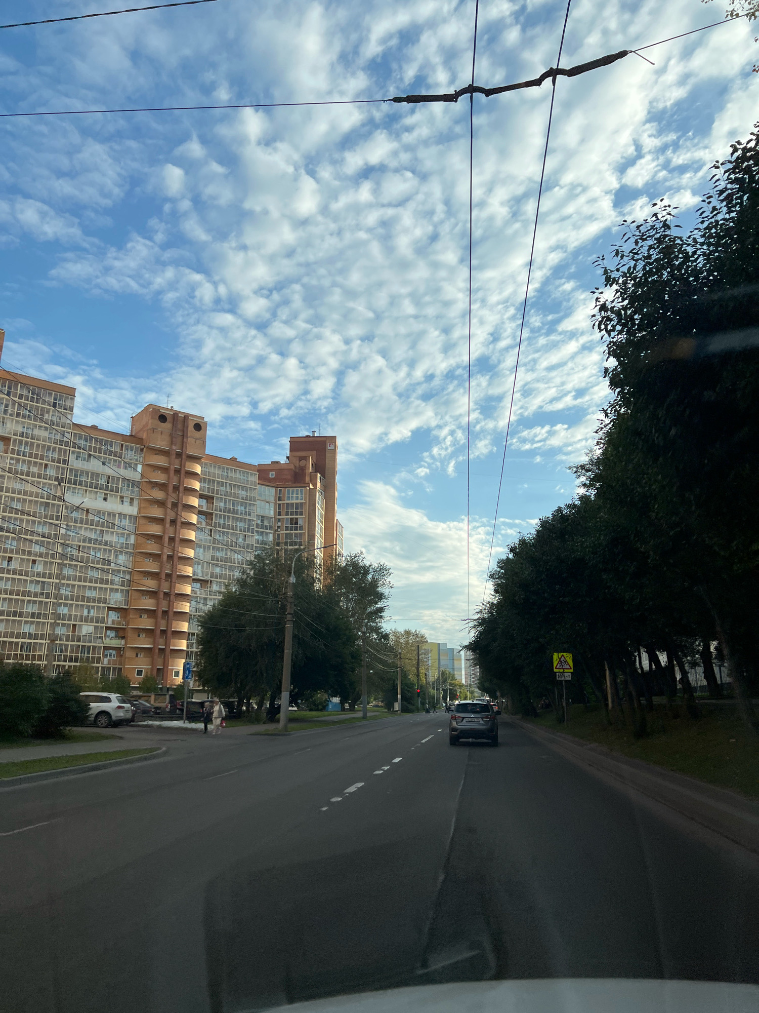 The image shows a wide, paved road in a city, likely Krasnoyarsk, Russia, with a car driving in the right lane and large, multi-story apartment buildings on the left side. The sky is blue with scattered white clouds, and overhead wires are visible. Trees line the right side of the road, and a pedestrian crossing sign is present. There are several parked cars and some pedestrians on the sidewalk. The apartment buildings are tall, with many windows and balconies. The overall impression is of a typical urban street scene on a partly cloudy day.