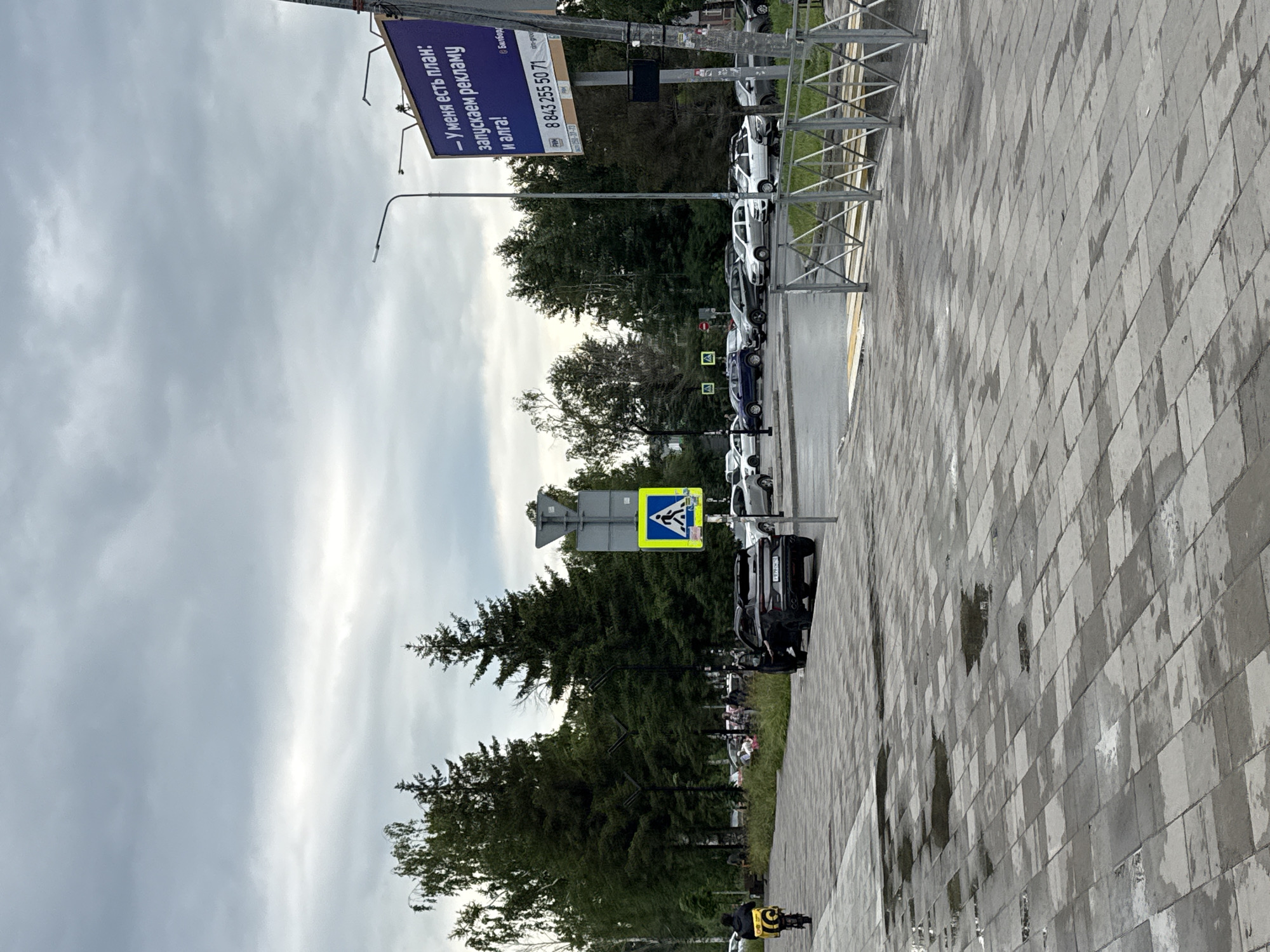 This is a medium shot of an outdoor scene in Kazan, Russia, on a cloudy day. The foreground is dominated by a wet, grey, paved walkway with scattered puddles, indicating recent rain. To the left, a person on a black electric scooter with a large yellow cargo bag is moving away from the viewer. In the middle ground, a black SUV with its driver's side door open is parked on the side of a road. Beyond the SUV, a pedestrian crossing is marked by a yellow and white striped crosswalk and a blue and white pedestrian crossing sign. Several cars are parked in a lot behind the pedestrian crossing, and a metal fence separates the walkway from the street. In the background, numerous tall green trees form a dense line, separating the urban area from what appears to be a sky filled with heavy, grey clouds. A large billboard with Russian text and a phone number is visible on the right side of the frame.