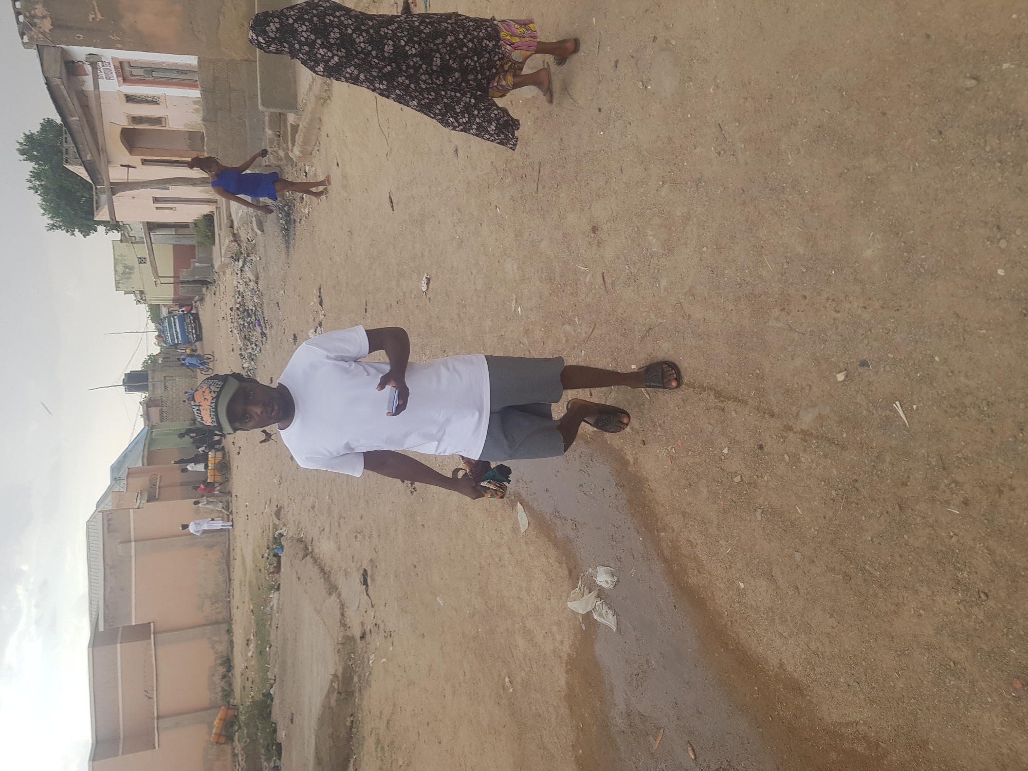 Captured in Dutsinma B, Nigeria, a man in a white shirt and gray shorts stands on a dirt road. He is holding a cell phone in his left hand and a wallet in his right. A woman in a dark floral dress walks behind him. A girl in a blue dress is running down the street, and there are buildings in the background. There is garbage in the street.