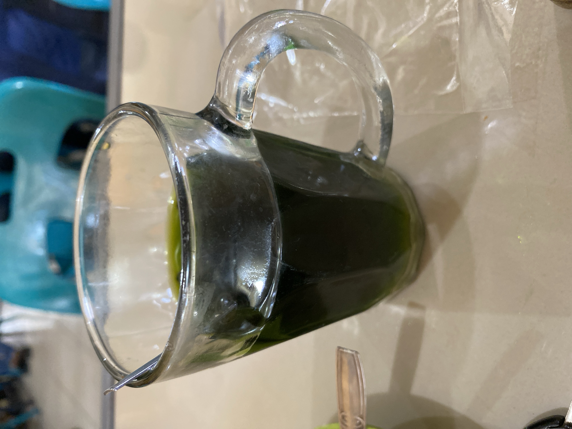 A close-up, eye-level shot shows a clear glass mug filled with a dark green liquid and a small silver spoon resting on the rim. The mug has a curved handle and sits on a light-colored, textured surface, possibly a table, which is partially covered by a clear plastic sheet. In the blurred background, hints of teal-colored chairs and furniture are visible, suggesting an indoor setting, likely a cafe or restaurant. The lighting is ambient and slightly diffused, creating a soft glow on the glass and liquid. The overall scene is still and suggests a moment of pause, possibly before or after consumption of the beverage. The image is focused on the mug and its contents, with the background elements intentionally out of focus. The location is Lhokseumawe, Indonesia.