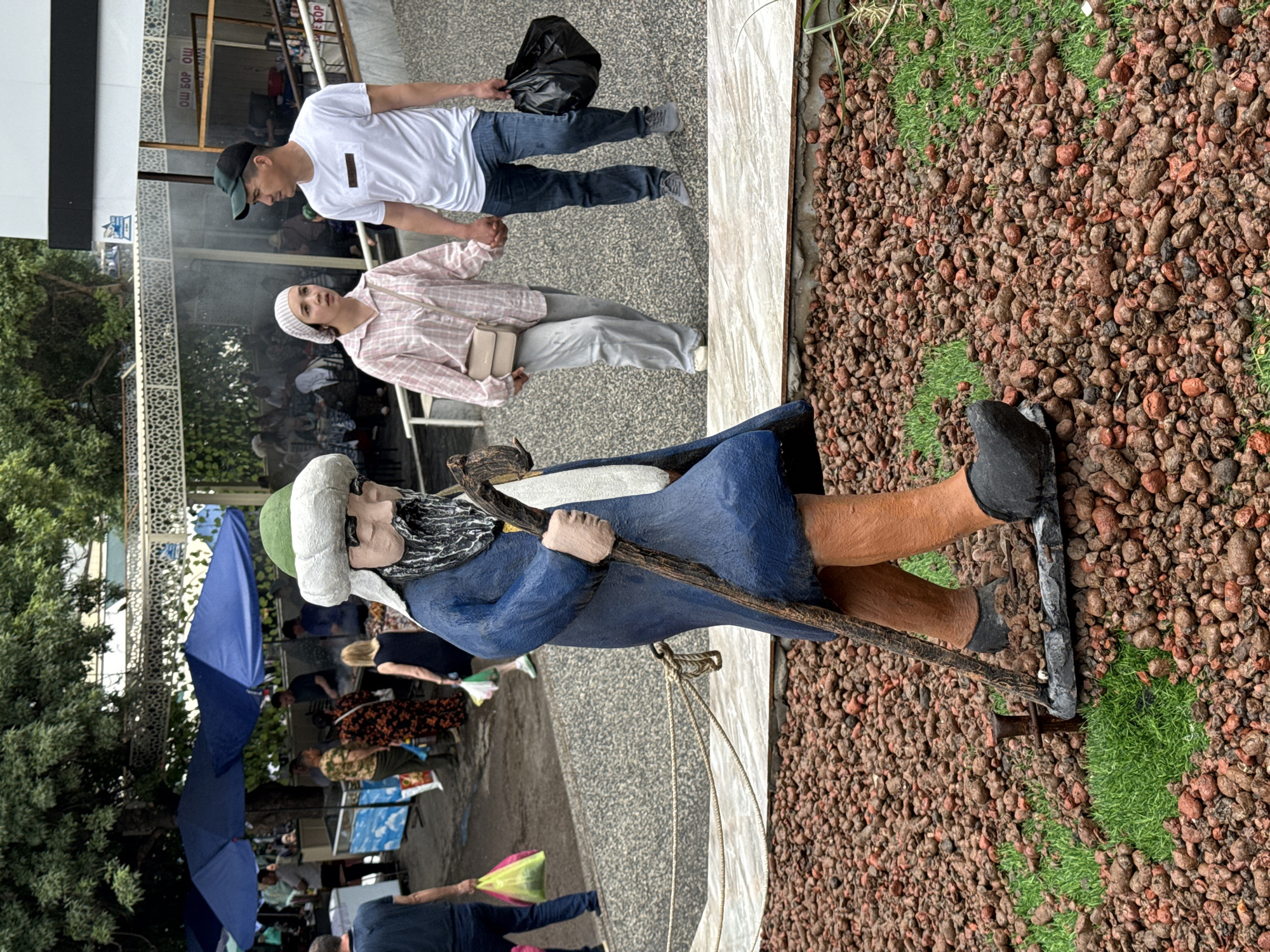 The image shows a view of Chorsu Bazaar in Tashkent, Uzbekistan. In the foreground, there is a statue of a man wearing a blue robe, a green and white hat, and holding a walking stick. The statue is positioned on a surface covered with small brown rocks. In the background, people are walking around and shopping. There are umbrellas shading them. A man and a woman are standing behind the statue, near a shop or restaurant. The man is wearing a white T-shirt and jeans, while the woman is wearing a pink plaid shirt and light-colored pants.