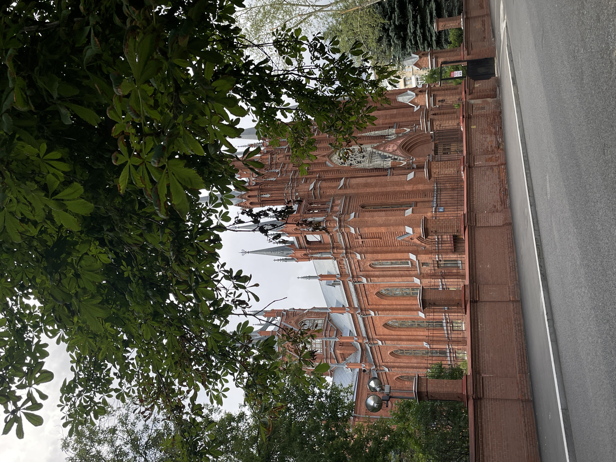 The image depicts the majestic Cathedral of the Immaculate Conception of the Holy Virgin Mary, a prominent Roman Catholic cathedral located in Moscow, Russia. The gothic-style architecture is characterized by its striking red brick facade, pointed arches, and numerous spires. The building is partially obscured by the lush green foliage of trees in the foreground, creating a framed view. A brick wall with decorative pillars and a black metal fence surrounds the cathedral, separating it from a paved street and sidewalk in the foreground. The sky is overcast, suggesting a cloudy day. No people are visible in the scene, and there are no discernible actions or interactions taking place. The environment appears to be urban, with the cathedral being the primary architectural subject.
