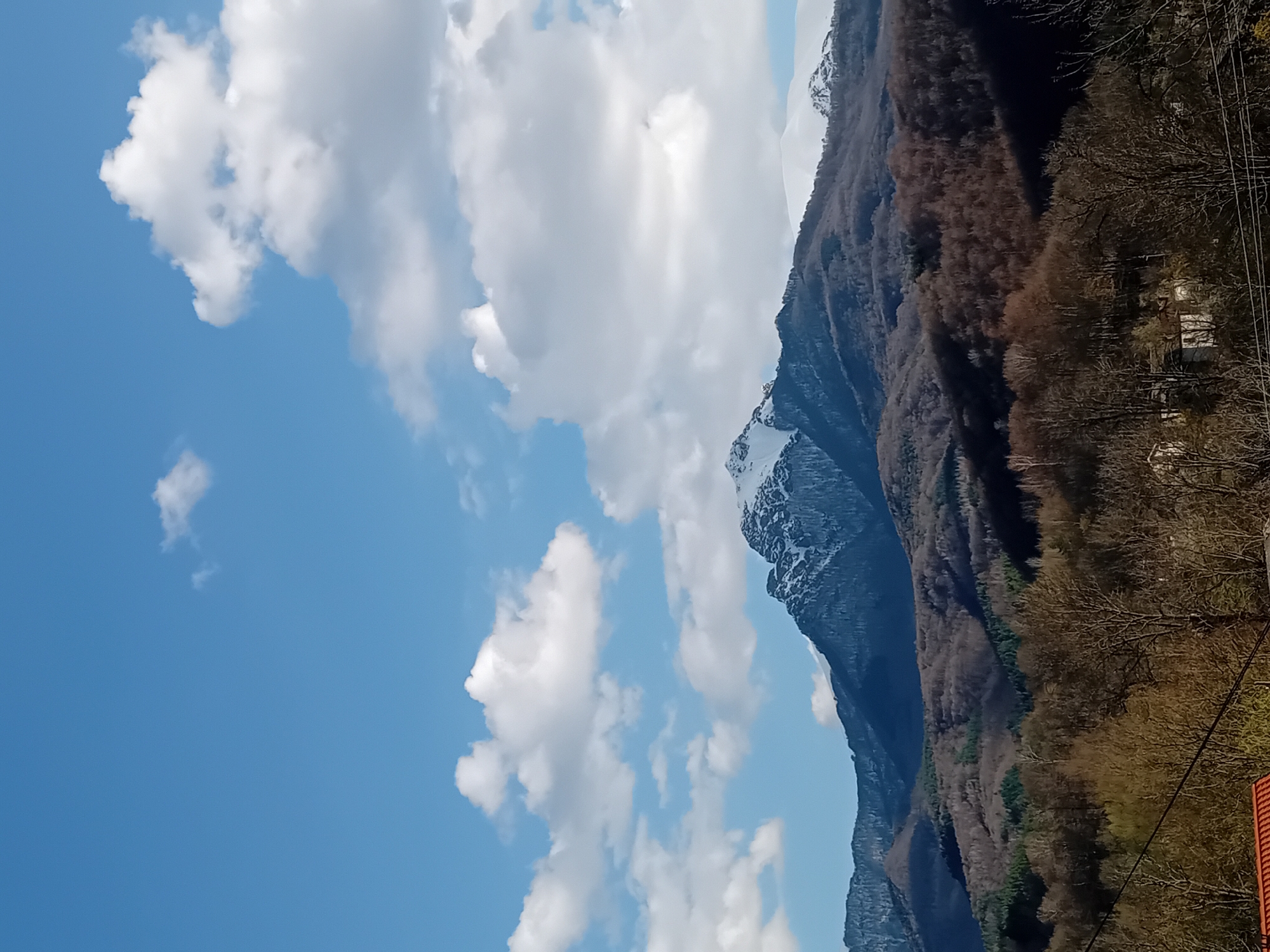 The image depicts a mountain range with snow-capped peaks under a partially cloudy sky. The lower slopes of the mountains are covered in trees, some of which appear to be bare. A portion of the sky is visible, with blue areas and fluffy white clouds. The image is vertical. The location is Unknown, Unknown.