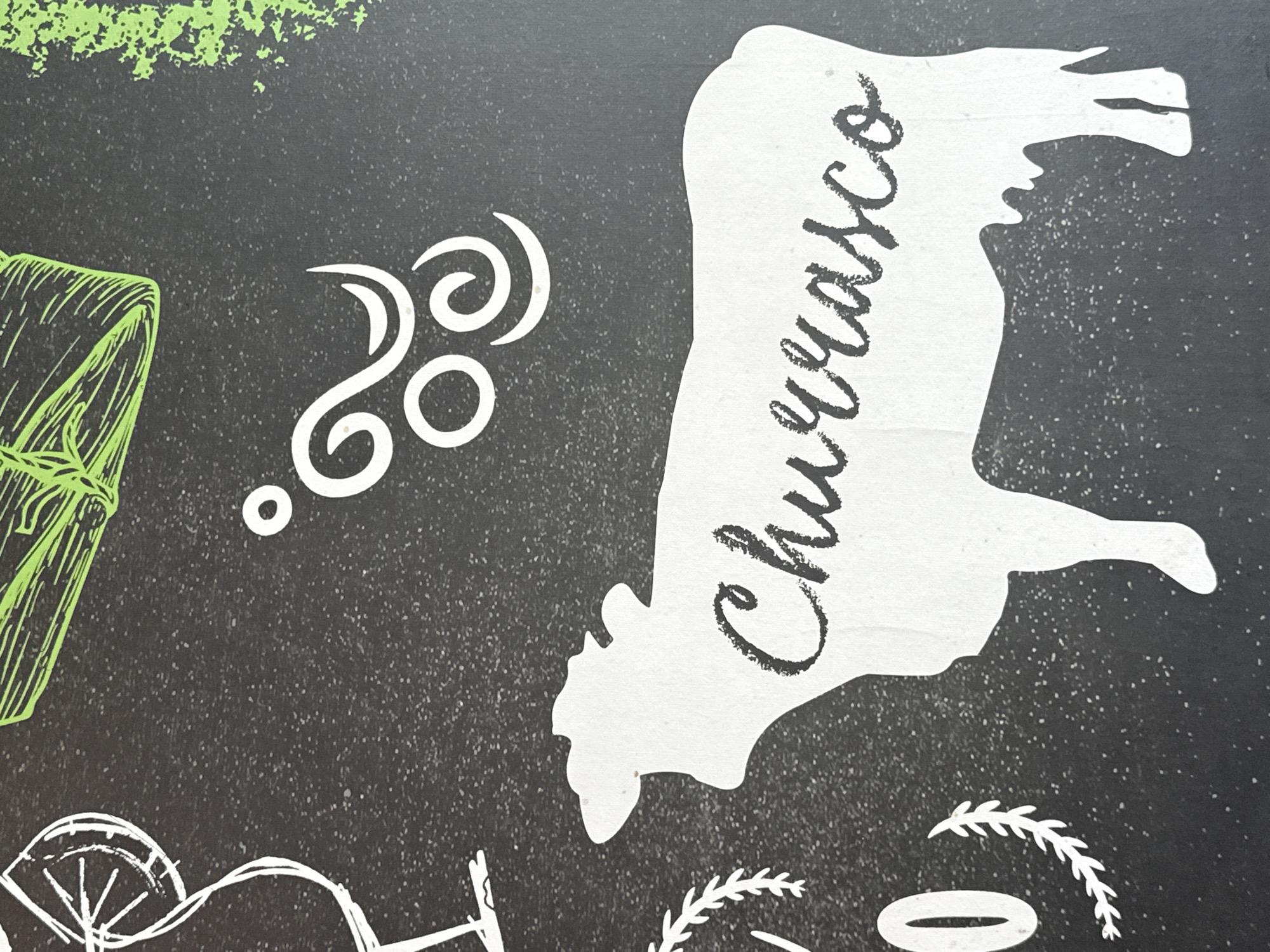 A chalkboard-style illustration features a white silhouette of a cow with the word "Churrasco" written across it in black script. The background is dark, speckled with white dots, resembling chalk dust. To the left of the cow are other white and green illustrations, including swirling decorative lines and what appears to be a wrapped bundle. Below the cow and other illustrations, partial drawings of what may be a cartwheel, wheat stalks, and an oval are visible. The style of the image suggests a promotional advertisement or menu design, possibly for a Churrascaria restaurant. The location is not discernible from the image.