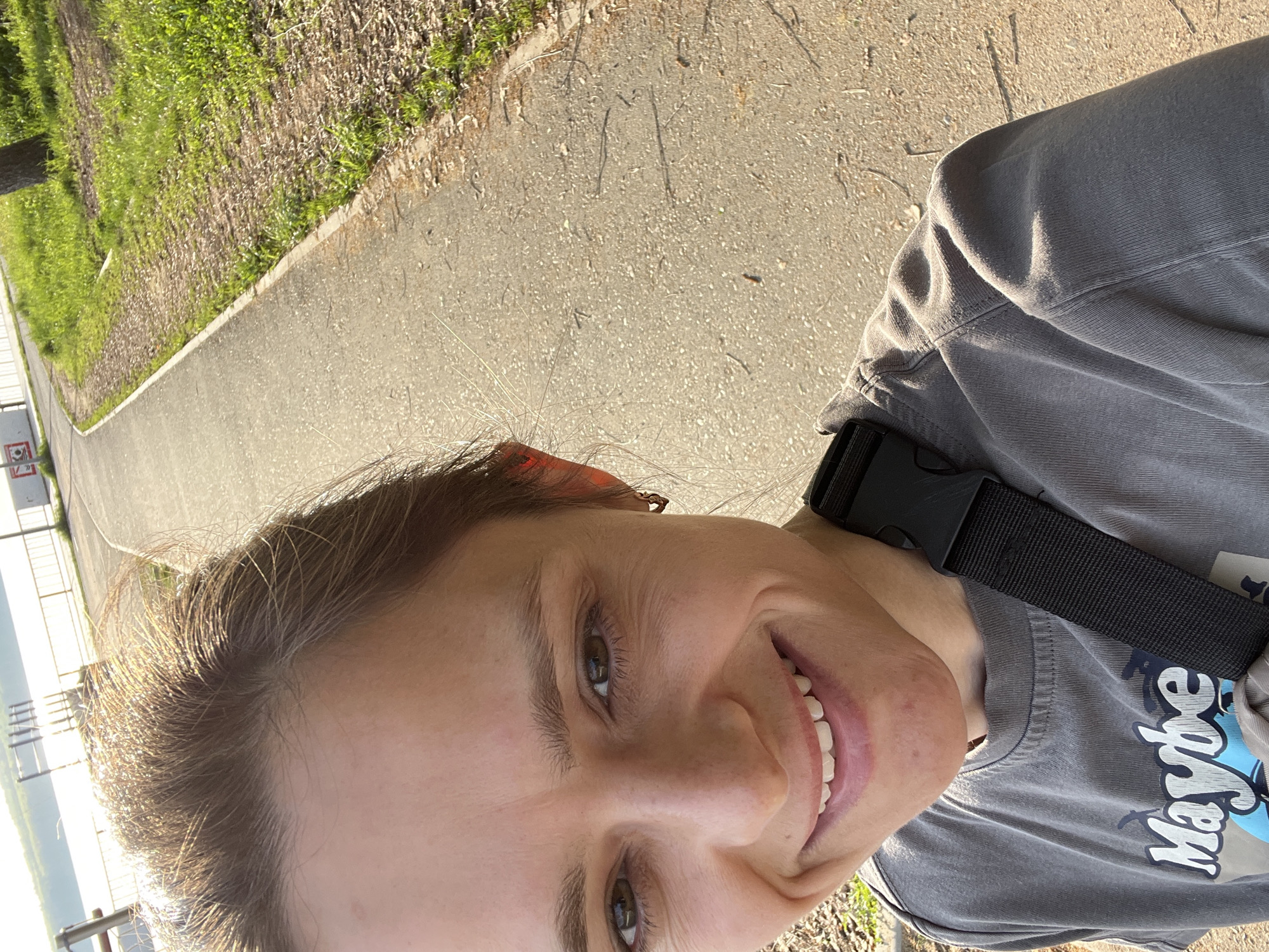 The image is a selfie taken by a woman. She is smiling at the camera. She is wearing a gray t-shirt with the word "Maybe" printed on it, and a black strap is visible across her chest. She is outdoors on a paved path. The background includes grass, a concrete barrier, and a distant body of water. The location is likely Chaikovsky, Russia.