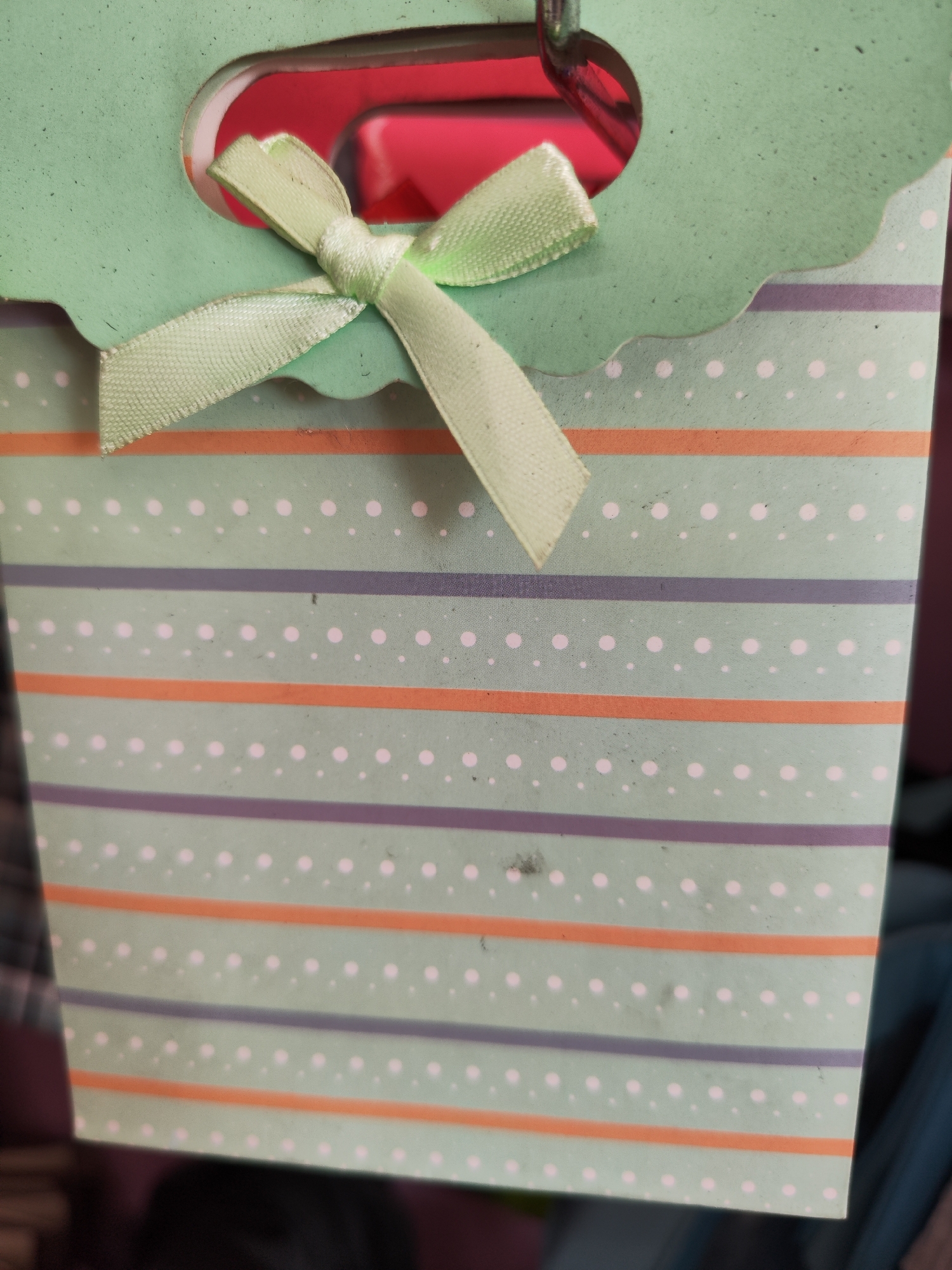A close-up, eye-level, medium shot captures the top portion of a light green paper gift bag with a scalloped edge. A light green ribbon is tied in a bow on the left side of the bag's opening, with one end of the ribbon extending downwards. The bag's surface is decorated with horizontal stripes of varying colors and patterns: a light purple stripe, followed by a thin orange stripe, and then a wider light green section with small white polka dots. This pattern repeats down the visible part of the bag. Inside the bag's opening, a bright pink object is visible, though its exact nature is unclear. The background is dark and out of focus, suggesting the bag is being held or is placed against a dark surface or in a dimly lit environment. The image does not contain any elements that would allow for identification of a specific location within Sragen, Indonesia.