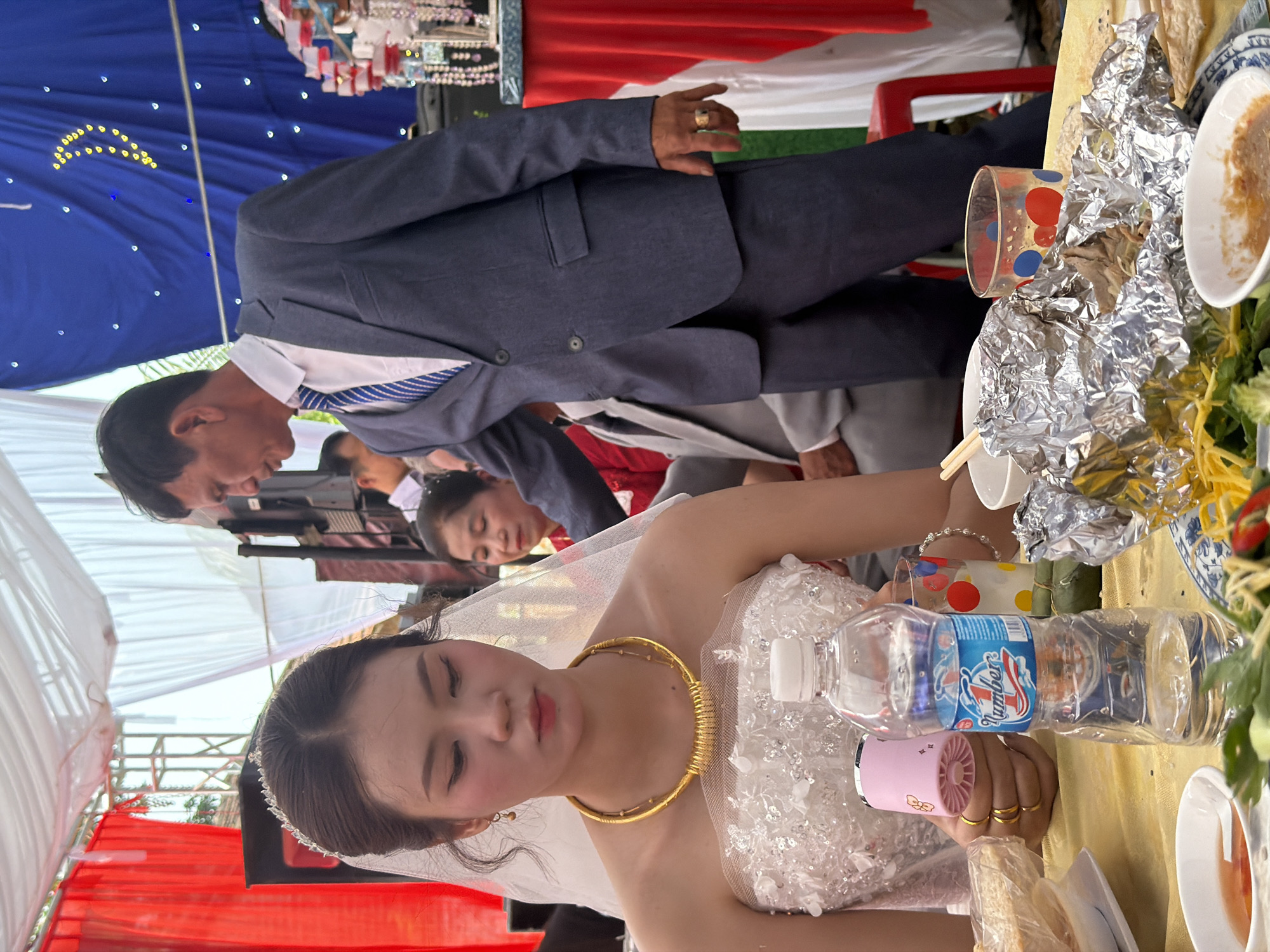 This image captures a daytime celebratory event, likely a wedding or engagement ceremony, set outdoors under a large tent or canopy. The scene is illuminated by bright, diffused light.
The primary subjects are a woman, presumably the bride, and a man, likely the groom. The bride, positioned in the foreground, wears a white, strapless or off-the-shoulder gown adorned with sequins or beads. She accessorizes with a large, golden choker necklace, a small jeweled tiara or headband, and gold rings on her right hand. She holds a small pink portable fan and a plastic water bottle. Her expression is serene. The groom, standing behind the bride, is dressed in a dark blue suit, a white collared shirt, and a blue and white striped tie. He wears a gold ring on his left hand.
Other individuals are visible in the background, including a woman in a red top and several partially obscured faces, suggesting a gathering of guests.
The table in the foreground is covered with a yellow tablecloth and features various dishes, some wrapped in aluminum foil, bowls, chopsticks, and a clear glass with red and blue polka dots. A plastic water bottle with a blue label, where the brand "Jumboz" (or similar) is partially visible, is also on the table.
The setting includes blue fabric draped overhead, decorated with yellow and white star-like embellishments, possibly forming a crescent moon. White canopy fabric is visible, along with red fabric draping in the background. A jewelry display stand with necklaces and bracelets is faintly seen in the far background.
Given the context of Tay Son Commune, Vietnam, the attire, decorations, and presence of specific food items (like a green wrapped item on the table, potentially a traditional cake) are consistent with a Vietnamese wedding celebration.
