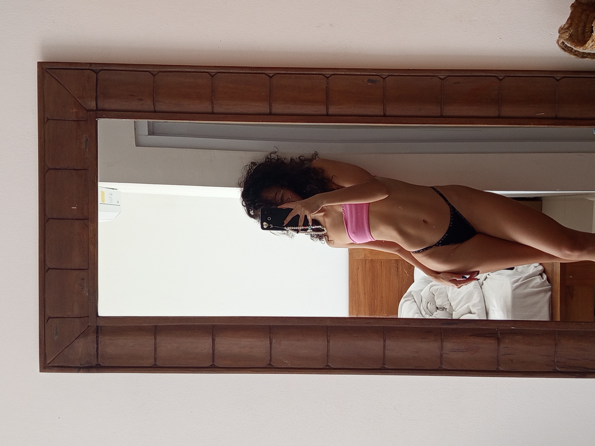 A mirror selfie of a woman in a pink top and black underwear is visible. The photo is taken in a room, likely in North Kuta, Indonesia, though no definitive landmarks confirming this are visible. The background includes a white wall, a wooden headboard, and rumpled white bedding. The woman is holding a smartphone in her left hand, obscuring part of her face.
