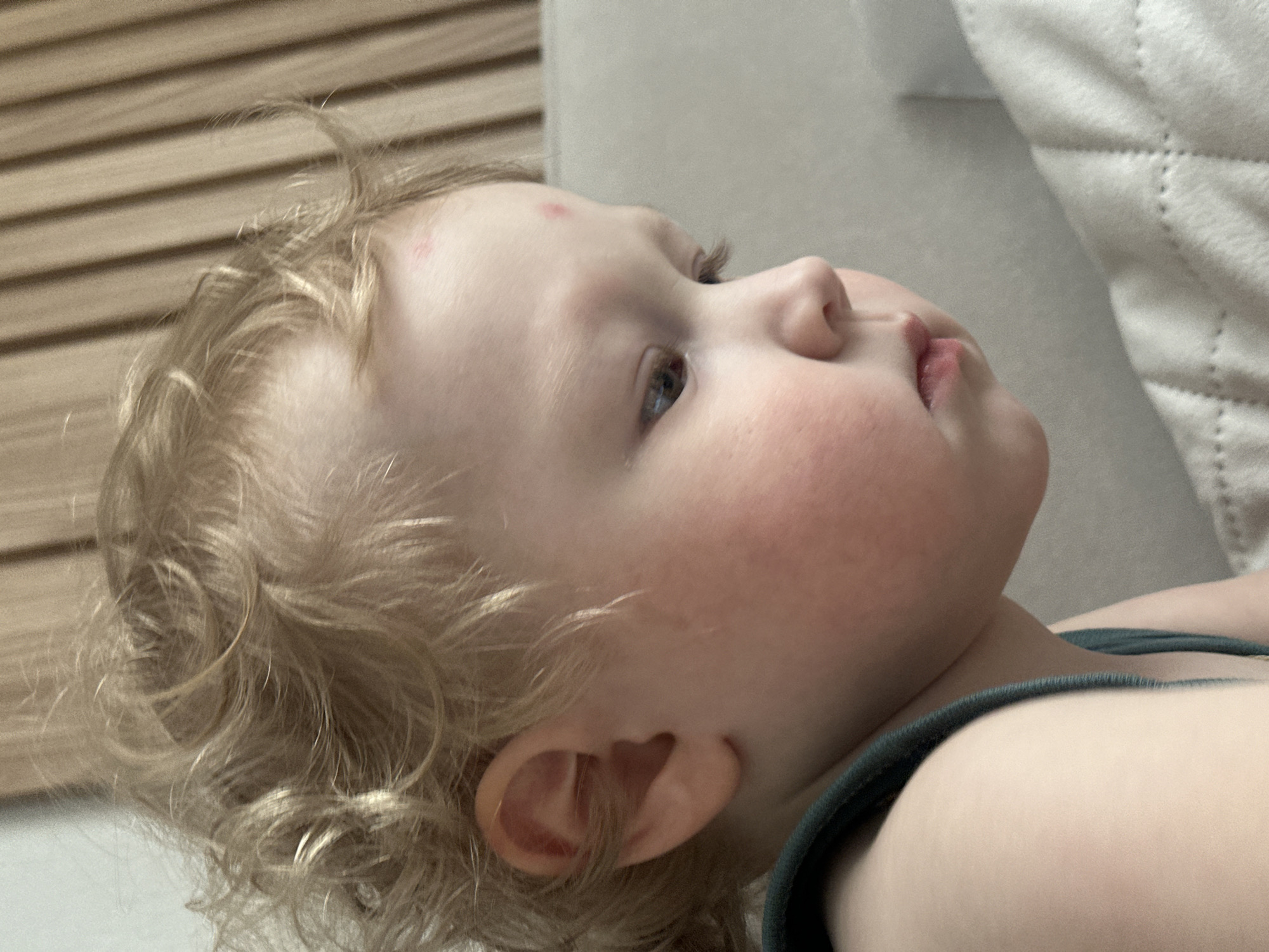 A close-up, medium-angle shot depicts a young child with blonde, curly hair lying on a soft, white surface. The child is facing upward and to the right of the frame, with their head resting on a cushion or pillow. Their right ear is visible, and there is a small red mark on their forehead. The child is wearing a dark green top. In the background are light-colored wooden slats and a quilted white surface. The location in Votkinsk, Russia is unknown.