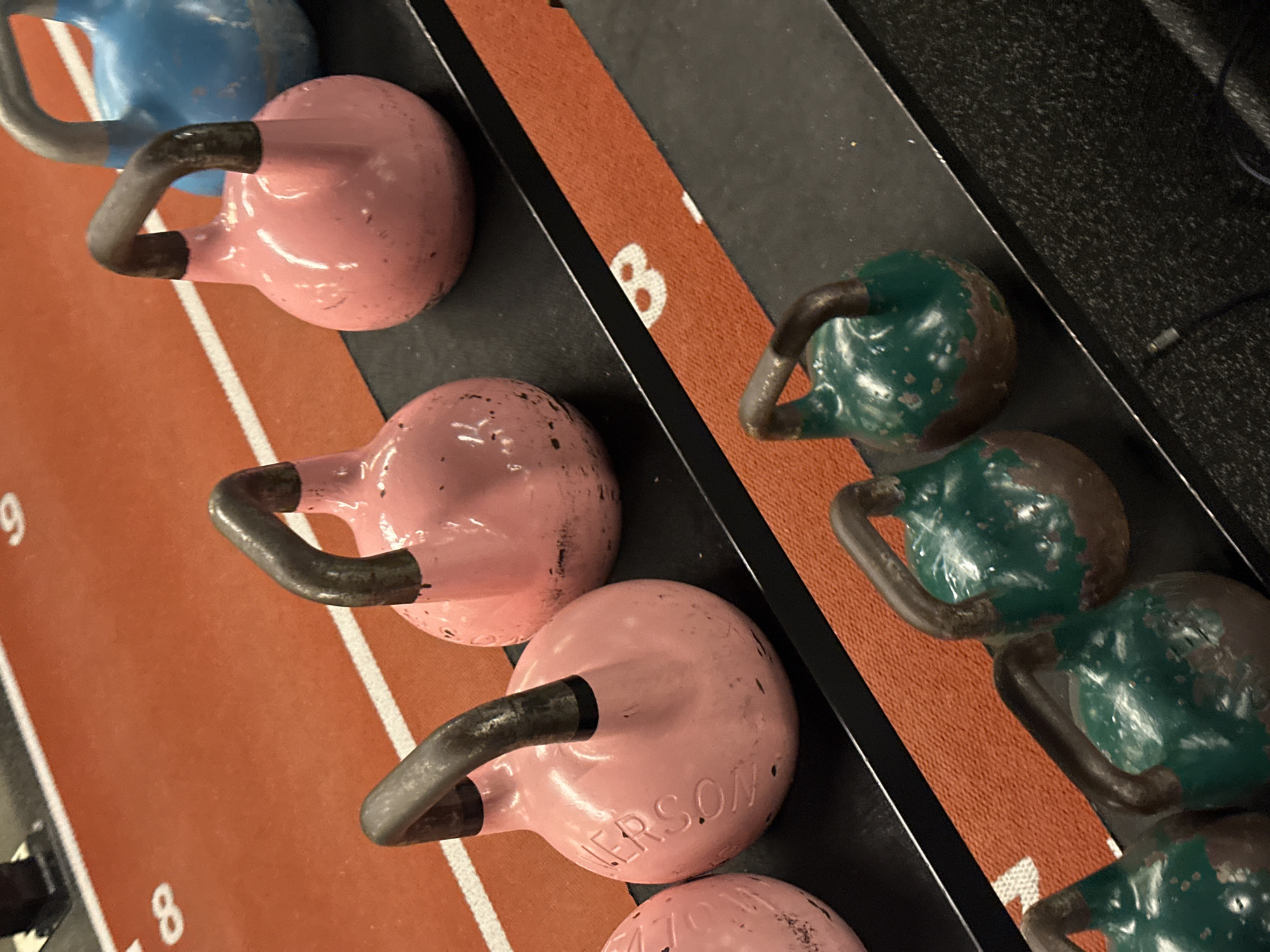 The image depicts a shelf with three rows of kettlebells in a gym setting. The kettlebells are arranged from left to right in the following colors: blue, pink, and green. The pink kettlebells have "PERSON" inscribed on one. An orange surface with white numbers runs perpendicular to the kettlebell rows. The number "8" and "9" are visible. The location of the scene is unknown.