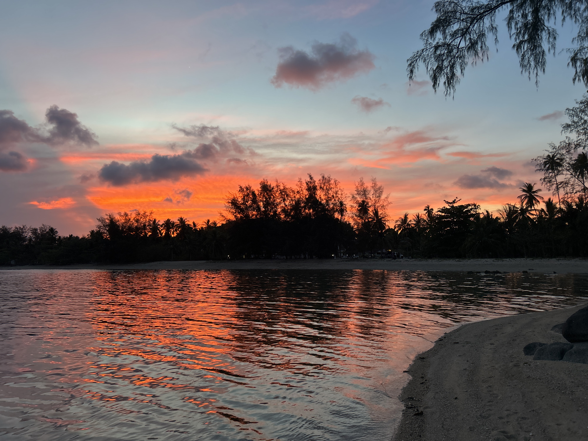 The image depicts a sunset over the water at Ban Bang Khao, Thailand. The sky is a mix of orange, pink, and blue hues with scattered clouds. Silhouetted trees line the horizon, reflecting in the water's surface. A sandy beach is visible in the foreground, with some rocks. There are no people visible in the scene. The overall environment is calm and peaceful.